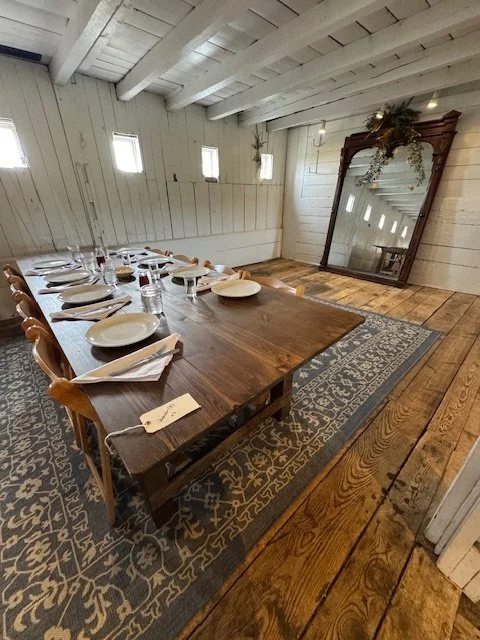 A rustic dining room with a long wooden table set with white plates, glasses, and napkins. There are wooden chairs around the table, and a large mirror leaning against the wall with greenery on top. The room features white wood-paneled walls, exposed