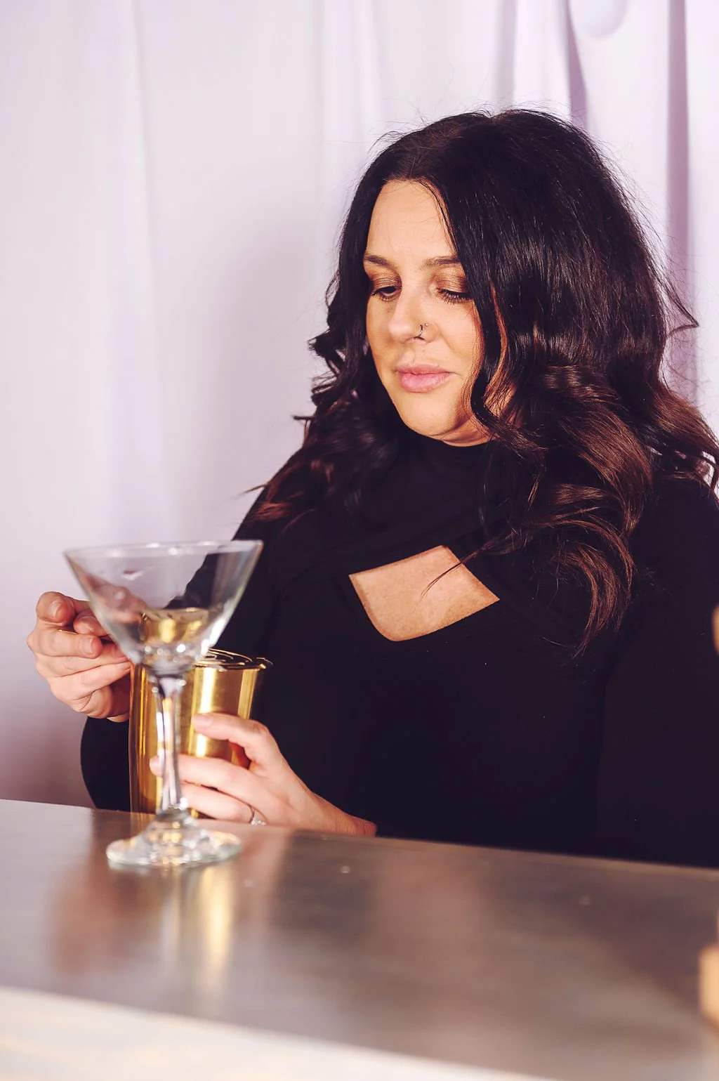 A woman with black wavy hair and a nose piercing is holding a martini glass and a gold-colored metallic cup, sitting in front of a white curtain.
