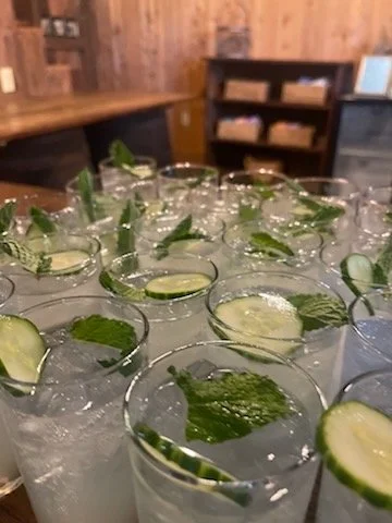Several glasses with drinks garnished with cucumber slices and mint leaves on a table, in a cozy wooden interior.