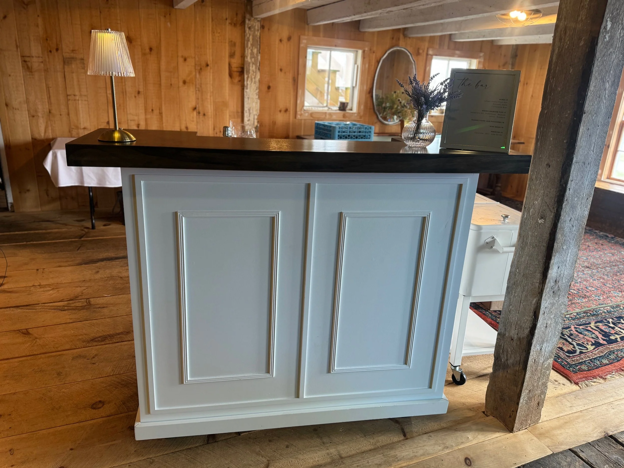A farmhouse-style bar with a dark wood countertop, white paneled front, and decorative molding, situated in a rustic wooden interior with exposed beams and wooden walls.