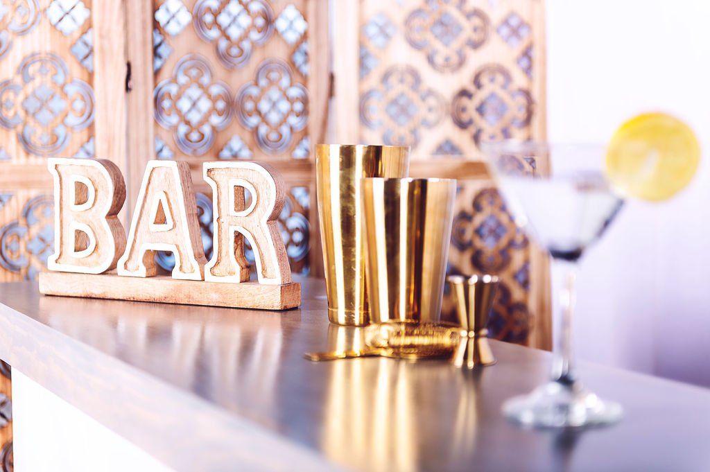 Bar setup with gold cocktail shakers, shot glasses, and an empty cocktail glass with lemon garnish, wooden 'BAR' sign, and decorative wooden wall with intricate cut-out patterns in the background.