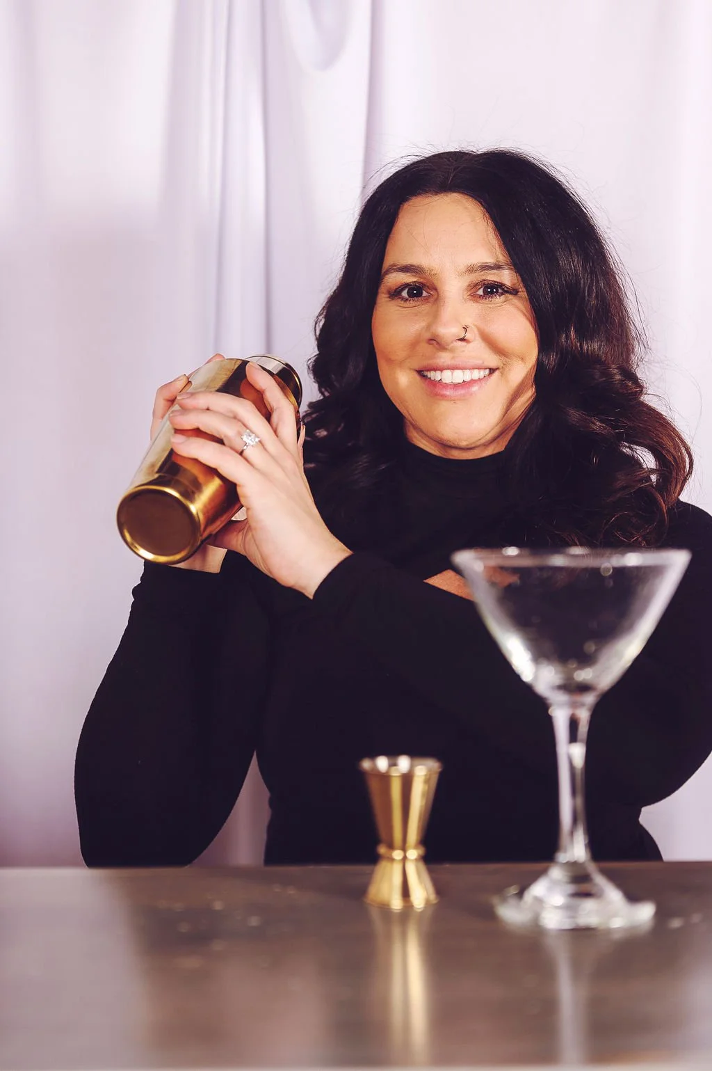 A smiling woman with long dark hair, wearing a black top, holds a cocktail shaker, standing behind a bar table with a martini glass and a gold jigger, against a light background.