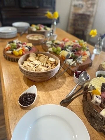 A wooden table set with various desserts, including cookies, fruit tarts with fresh fruit and whipped cream, a chocolate cake decorated with fruit, and a small dish of jam or preserves. There are also dishes, a vase with yellow flowers, and serving u