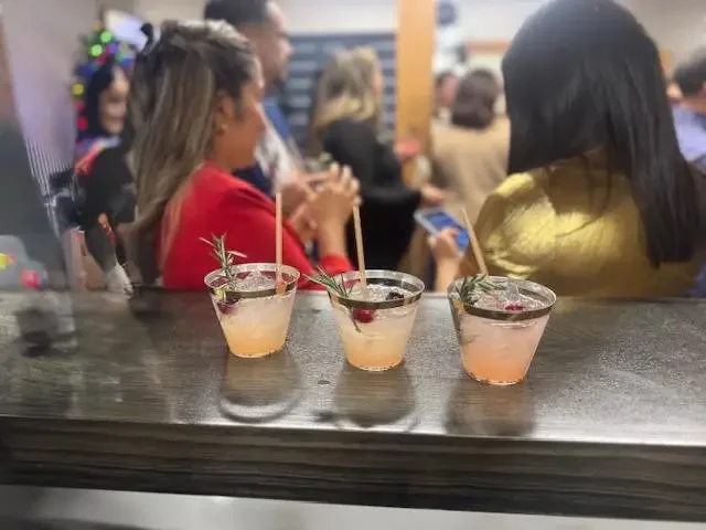 Three cocktails garnished with cherries and sprigs of herbs on a bar counter, with people socializing in the background.