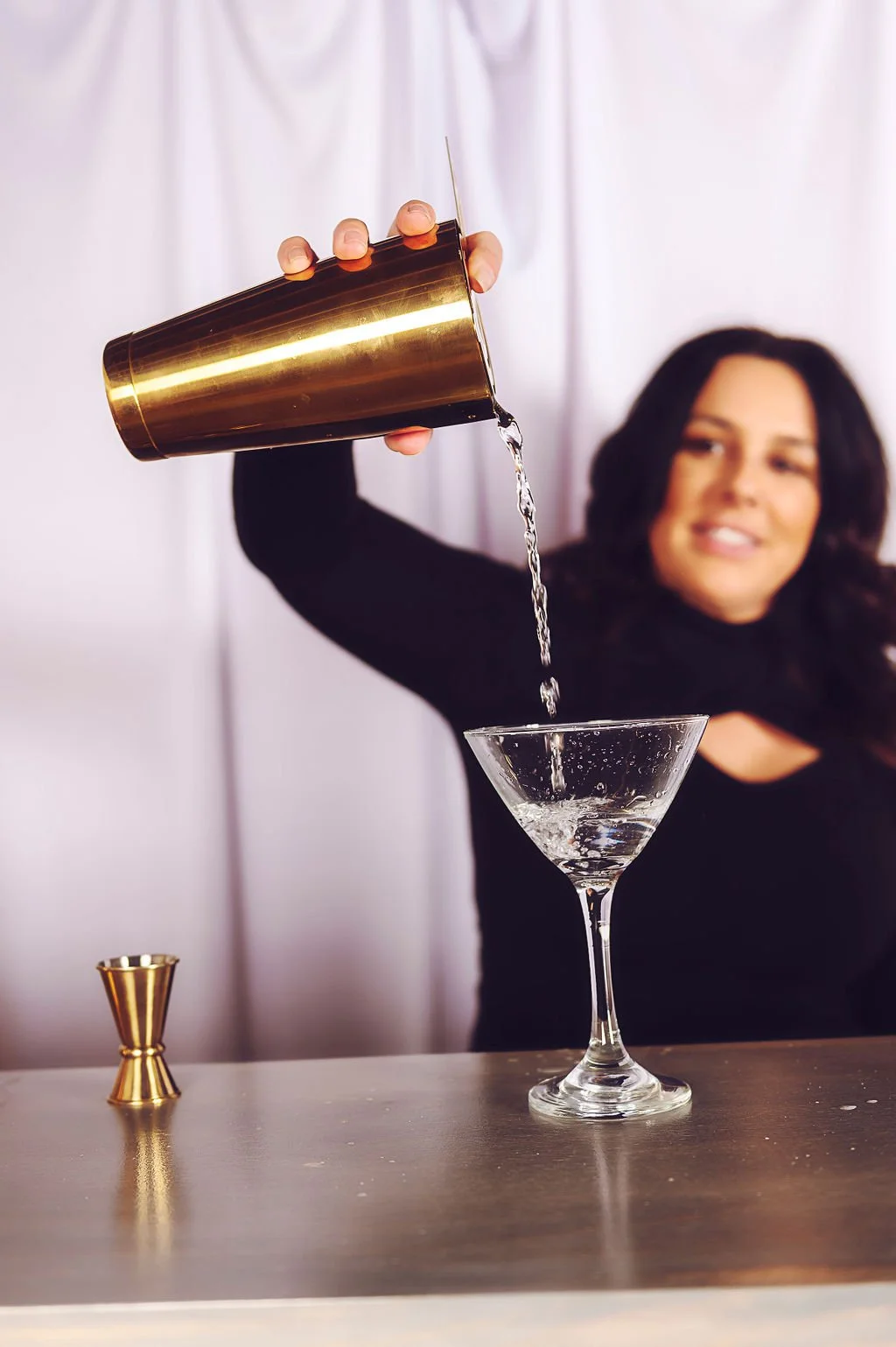 Woman pouring liquid from a gold shaker into a martini glass on a bar countertop, with a black jigger next to it and a white backdrop.