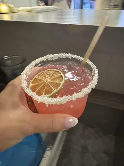 A pink cocktail in a glass with a salted rim, garnished with a lemon slice, and a straw.
