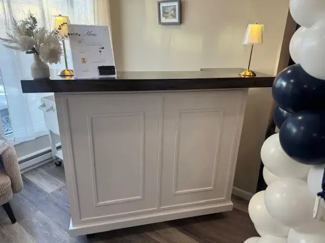 A white reception desk with a dark wood top, decorated with a white vase of flowers, two gold lamps, and a small menu stand, situated in a room with gray flooring, a window with sheer curtains, and a cluster of blue, white, and black balloons on the 