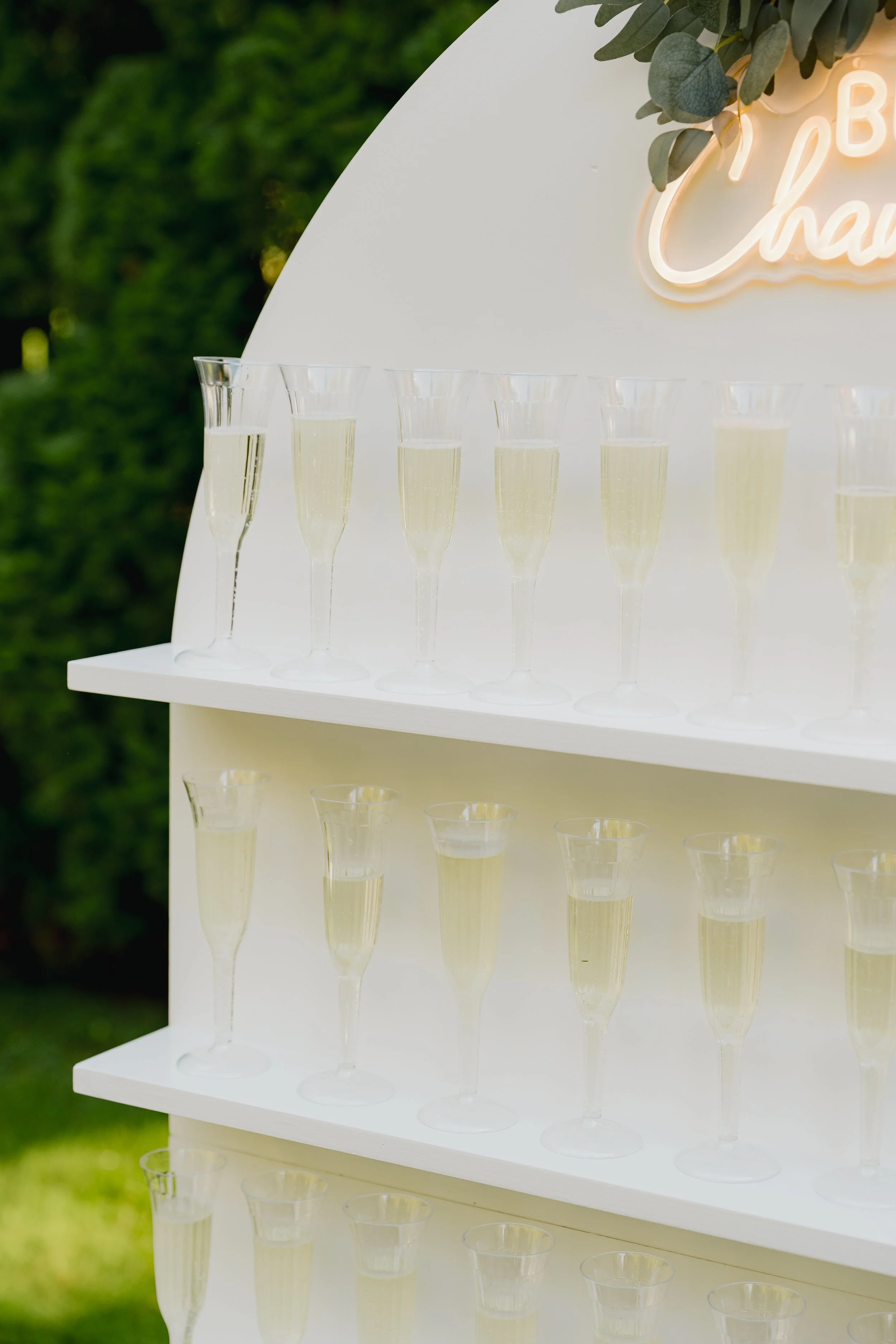 Multiple flutes of champagne on white shelves, outdoors, with greenery in the background and a neon sign that says 'Cheers' in cursive.
