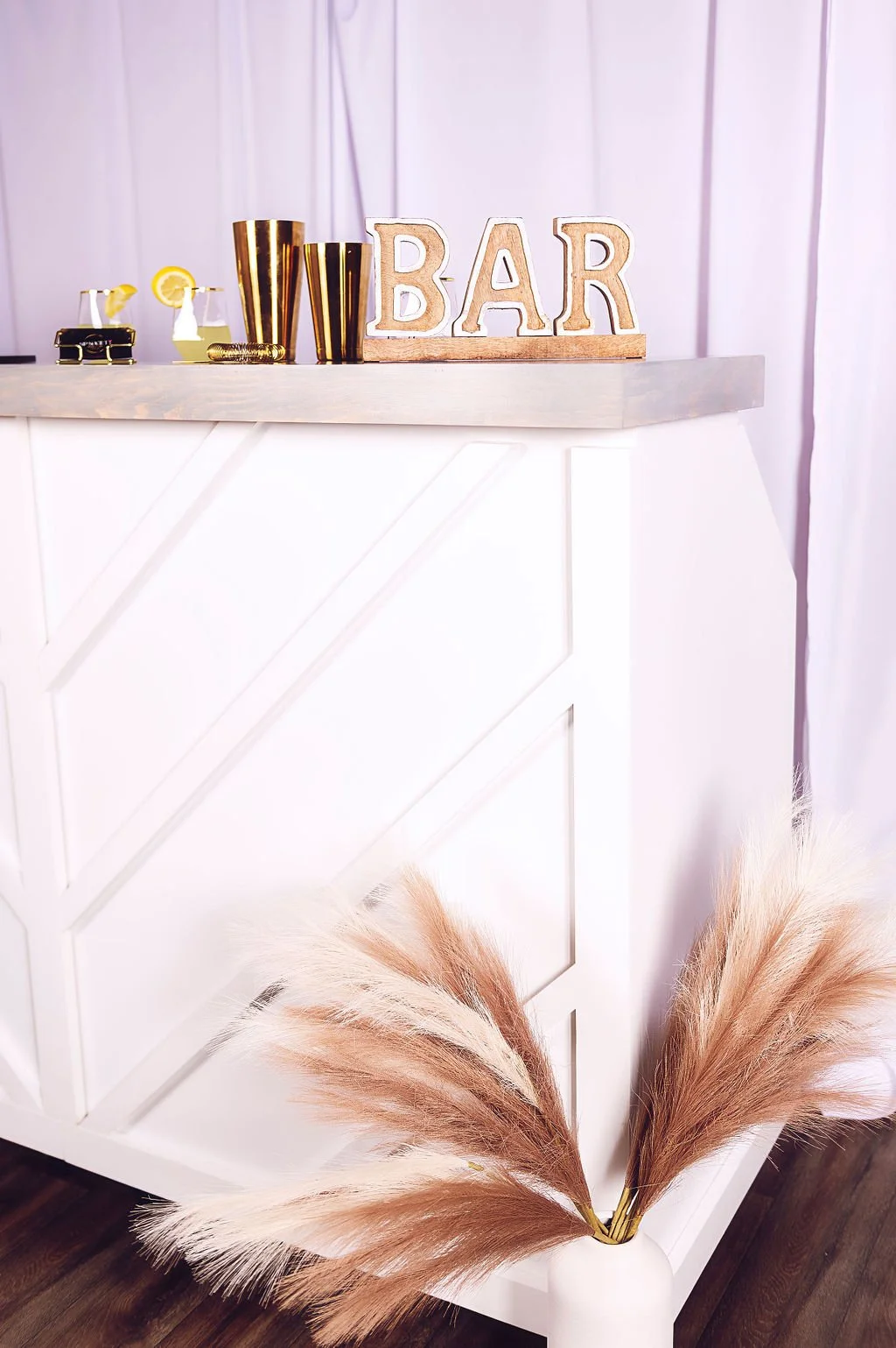 Decorative bar area with gold and black glasses, a gold bracelet, and a sign spelling 'BAR' on a white counter adorned with pampas grass in a white vase.
