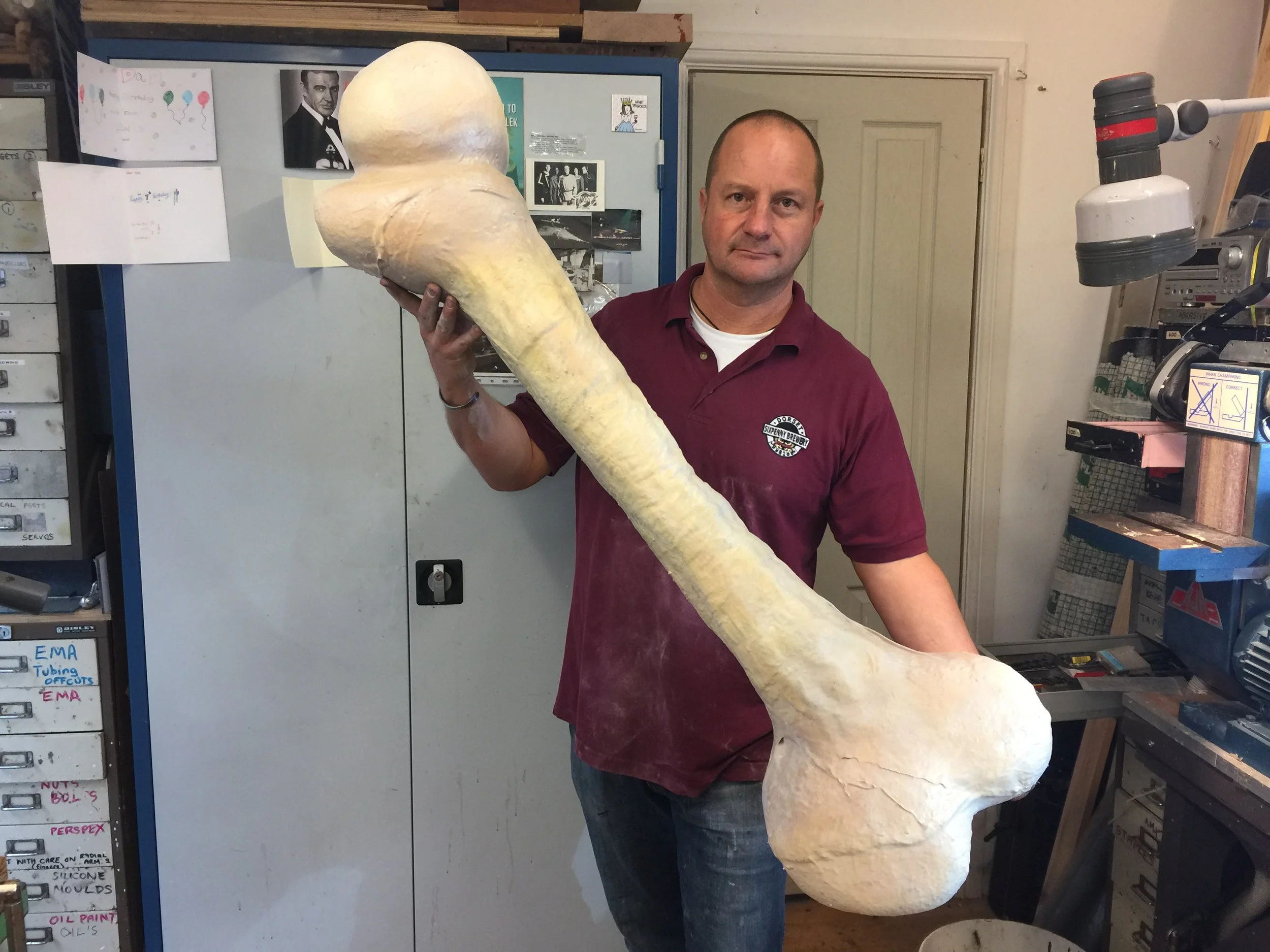 A man holding a large, oversized replica of a bone in a workshop or studio.