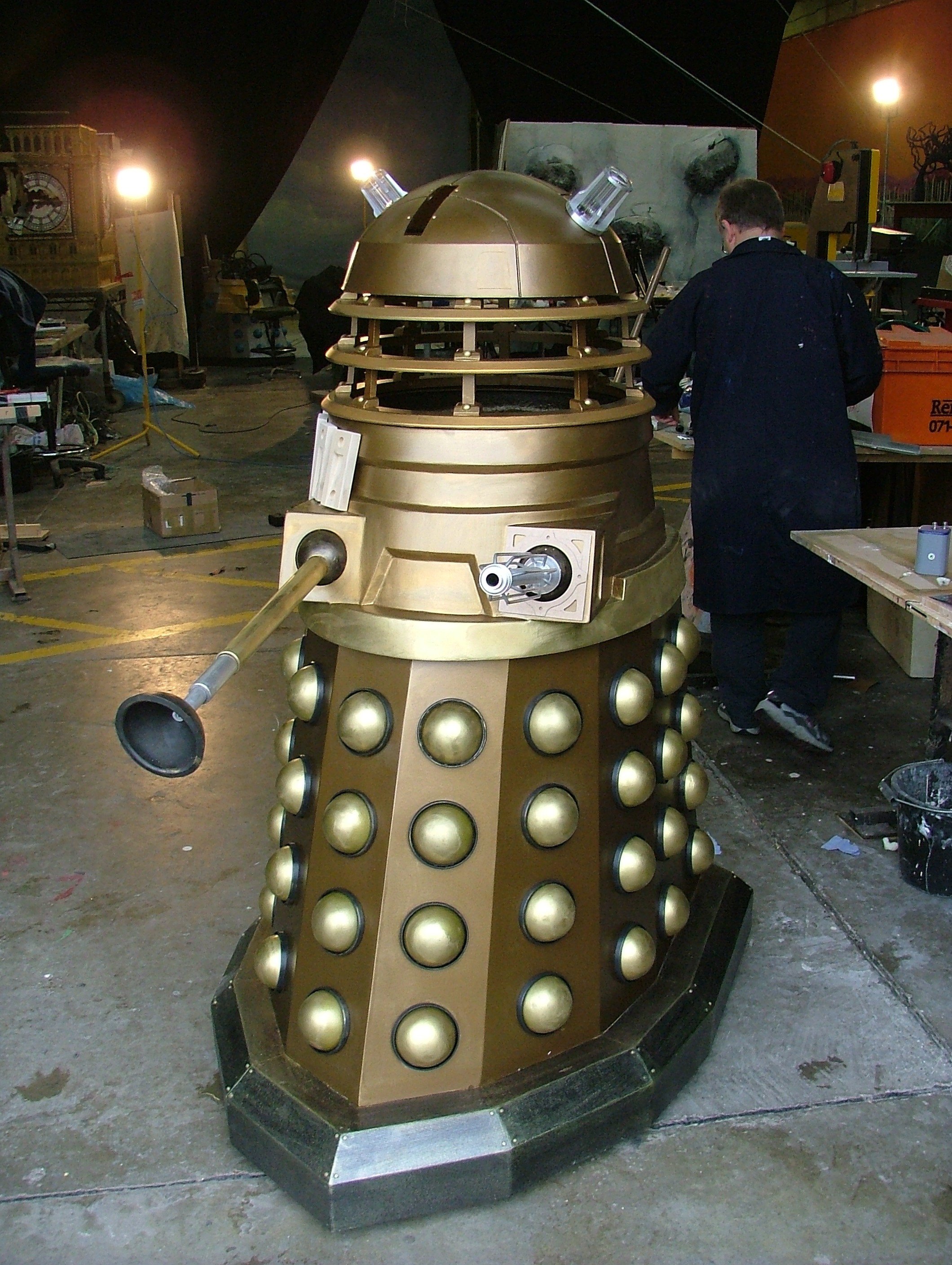 A life-sized Dalek replica from Doctor Who, painted in gold, standing in a workshop with a person working nearby.