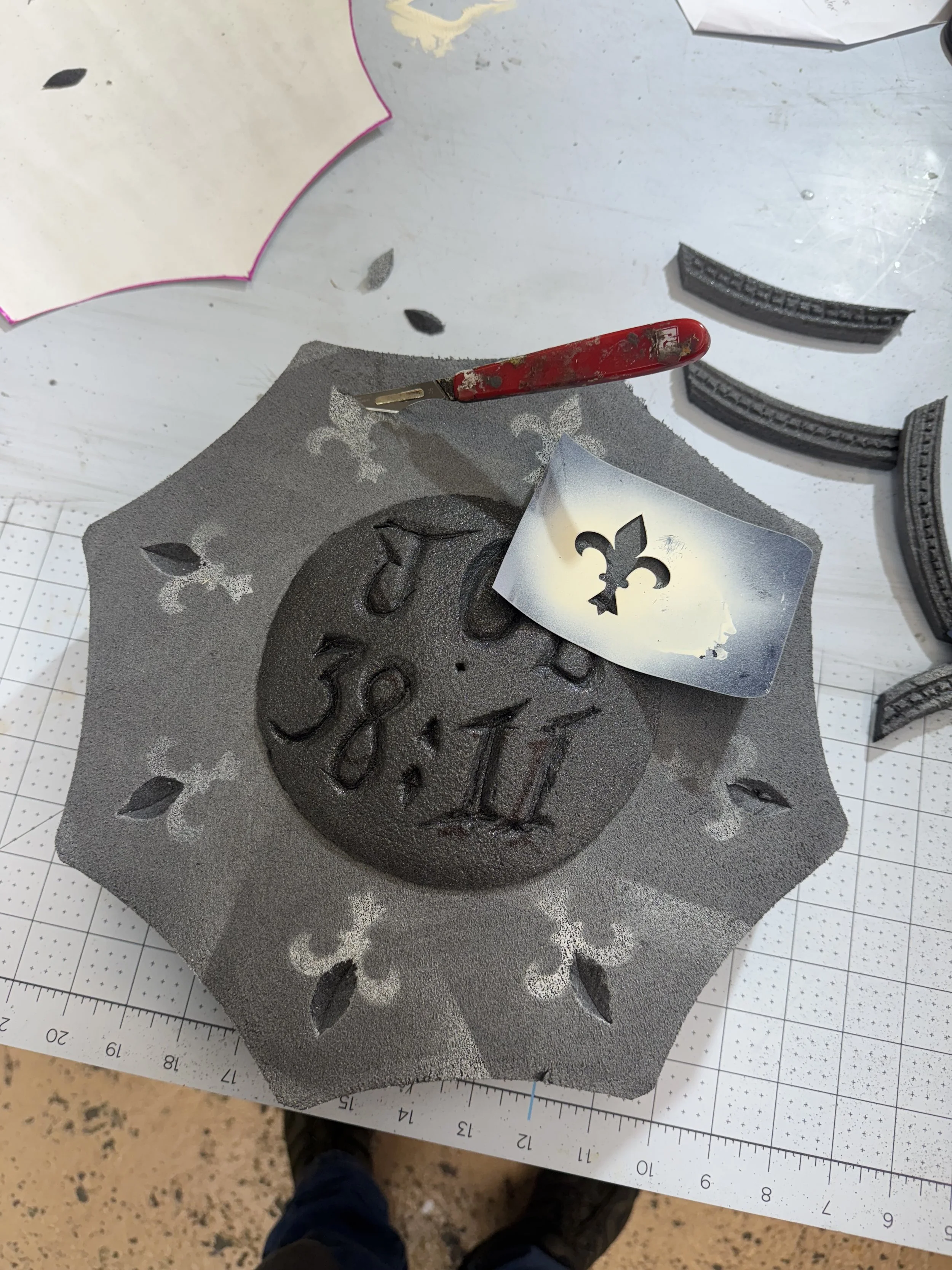 A black foam craft with a date carved in it, surrounded by a decorative hole-punched cutout. A stencil stencil with a fleur-de-lis symbol is placed on top, and a red craft knife is resting on the foam. There are additional foam strips and paper cutou