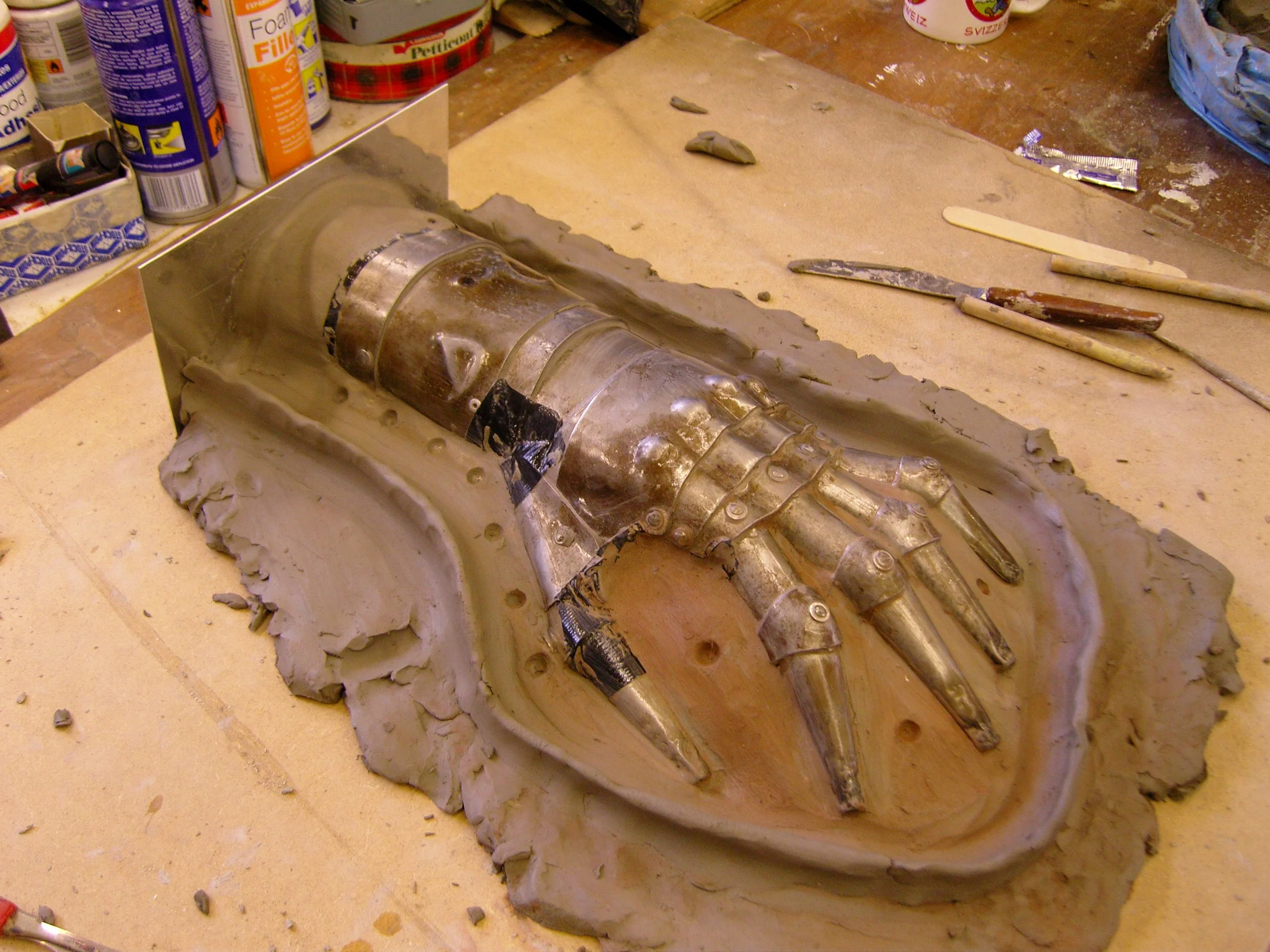 A detailed sculpture of a robotic or biomechanical hand, made from metal and clay materials, lying on a worktable surrounded by sculpting tools and supplies.