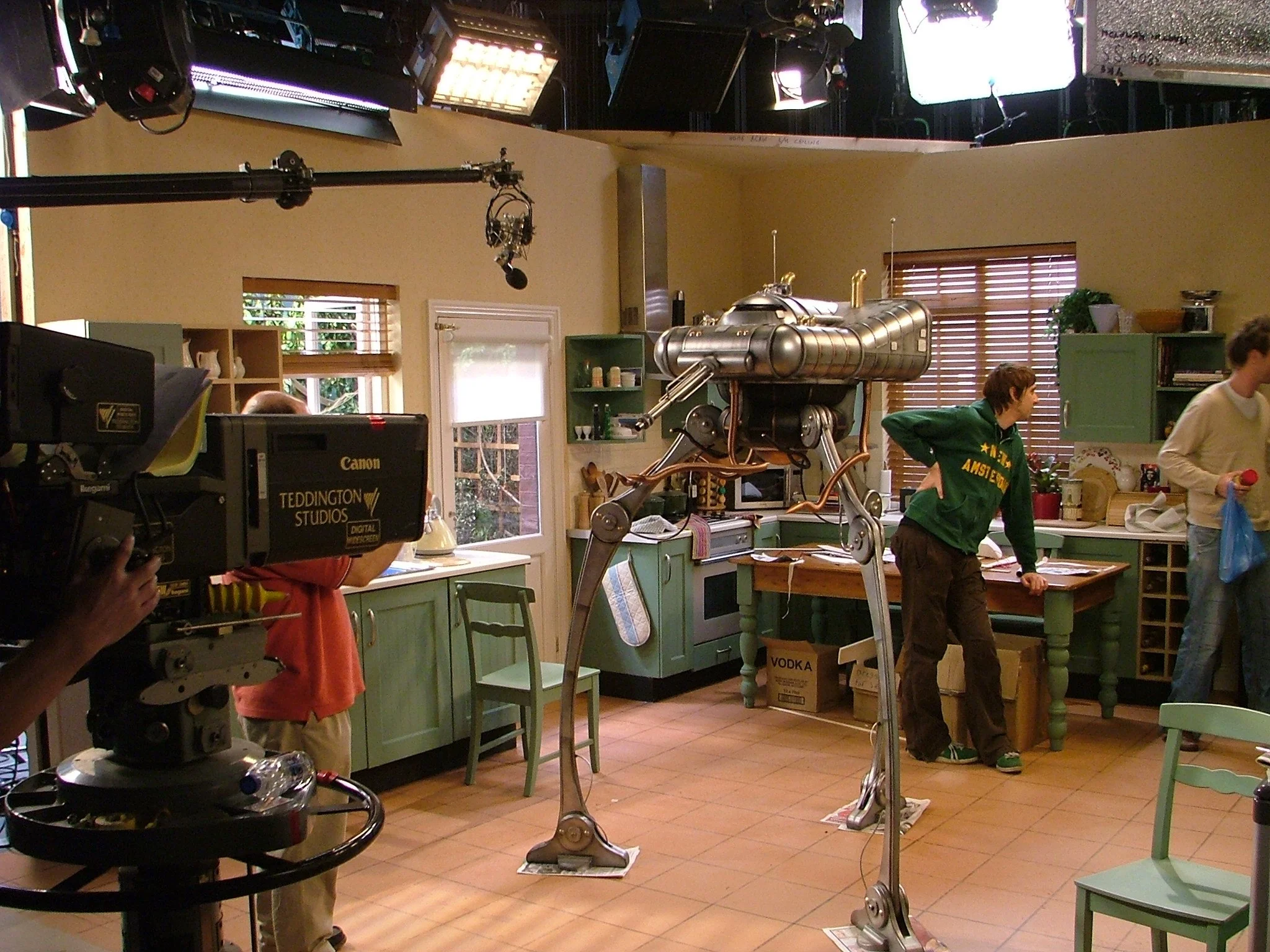 A television or film set designed to look like a kitchen with green cabinets and wooden countertops. A large robotic prop resembling a mechanical spider or alien creature stands on the kitchen floor. Crew members and actors are present, including a p