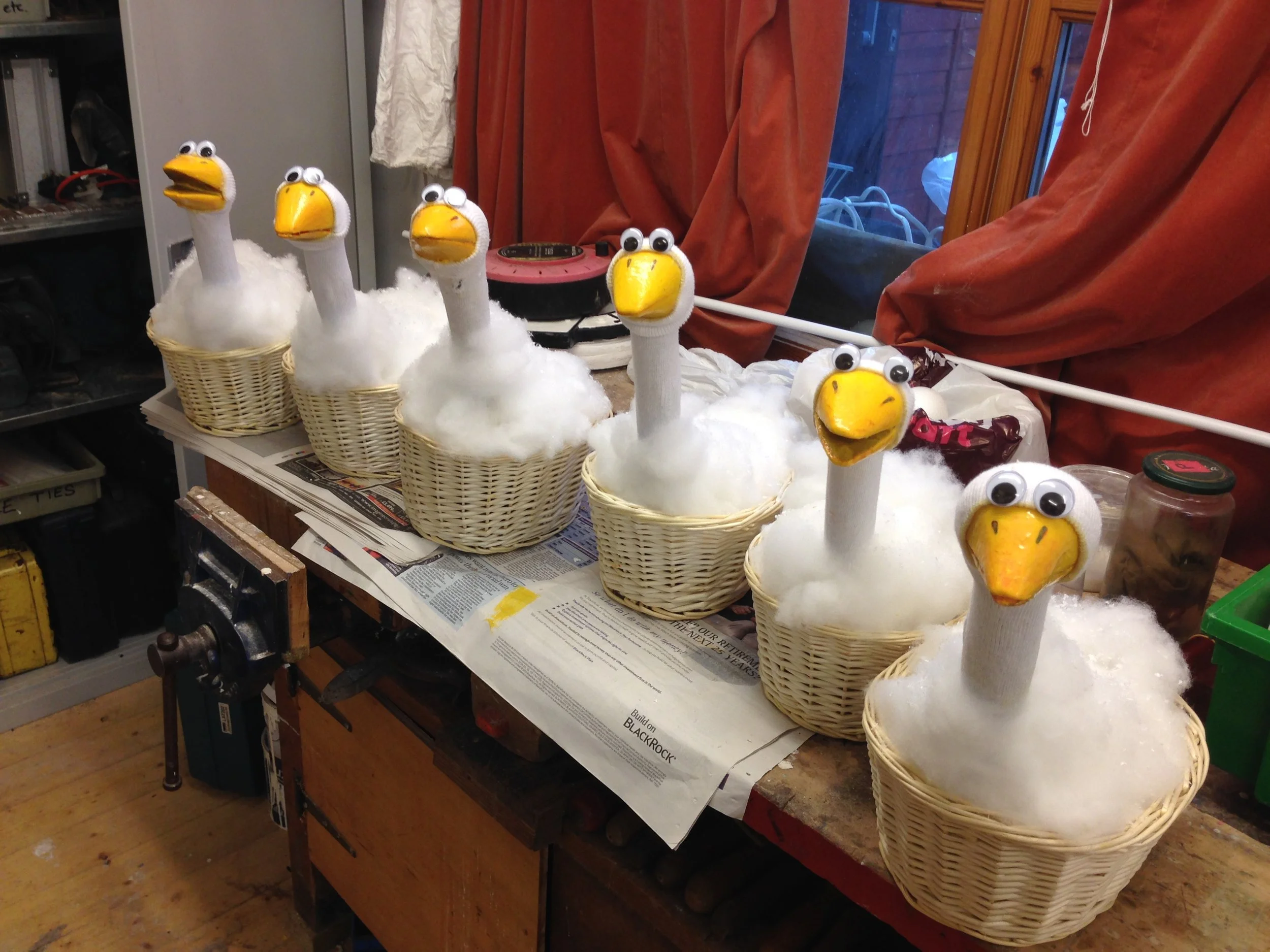 Seven craft goose figures made with baskets, cotton stuffing, and sock heads with googly eyes and yellow beak shapes, arranged on a workbench in a workshop.