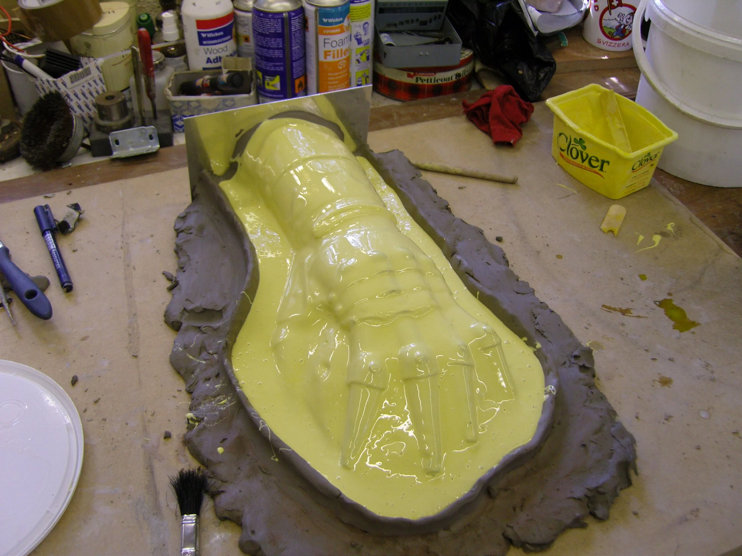 A workbench with an art project in progress, mold of Darth Vader's mask filled with yellow resin, surrounded by tools, paints, and supplies.