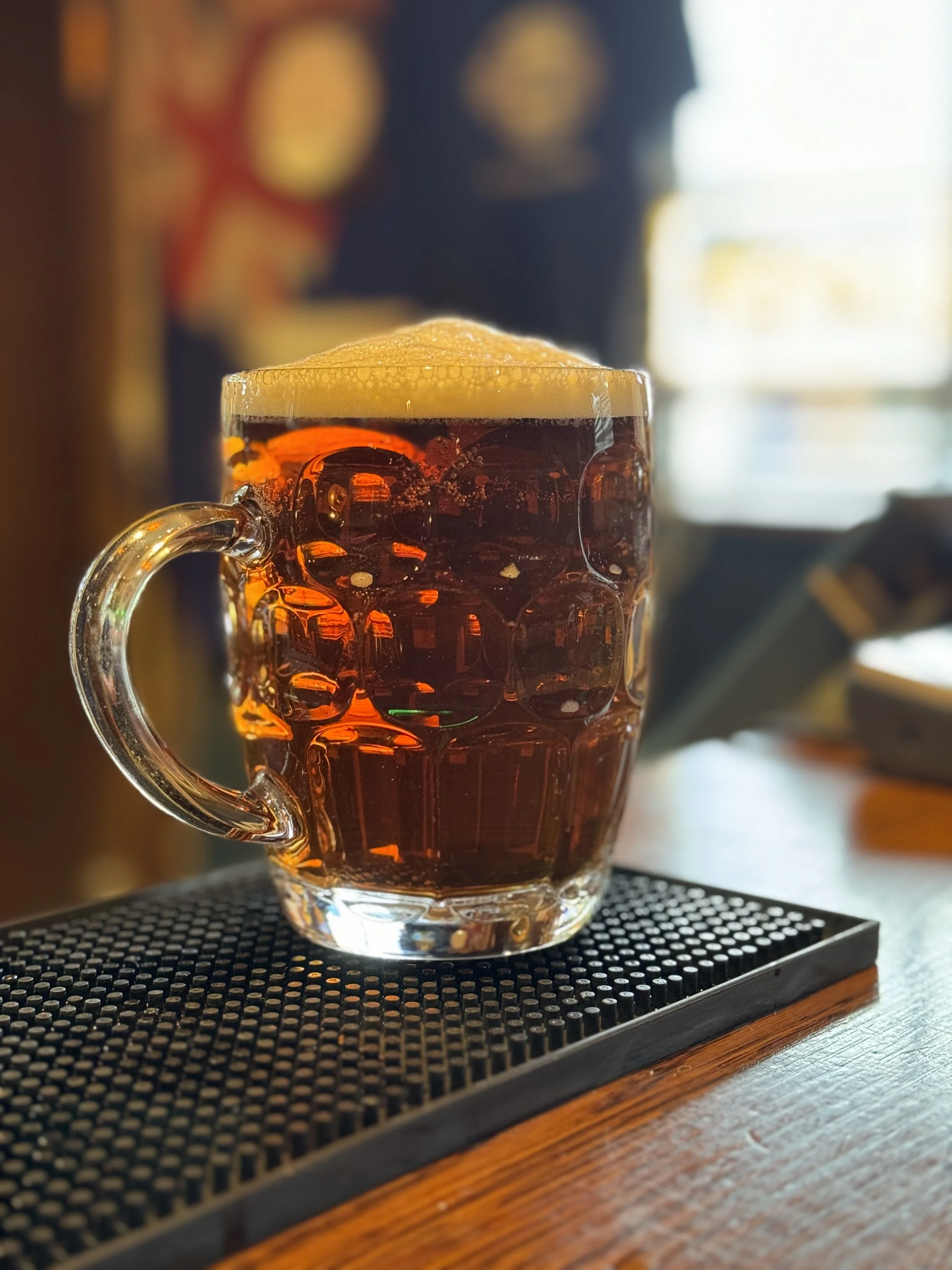 A mug of dark beer with a foamy head sitting on a black textured bar mat on a wooden bar counter.