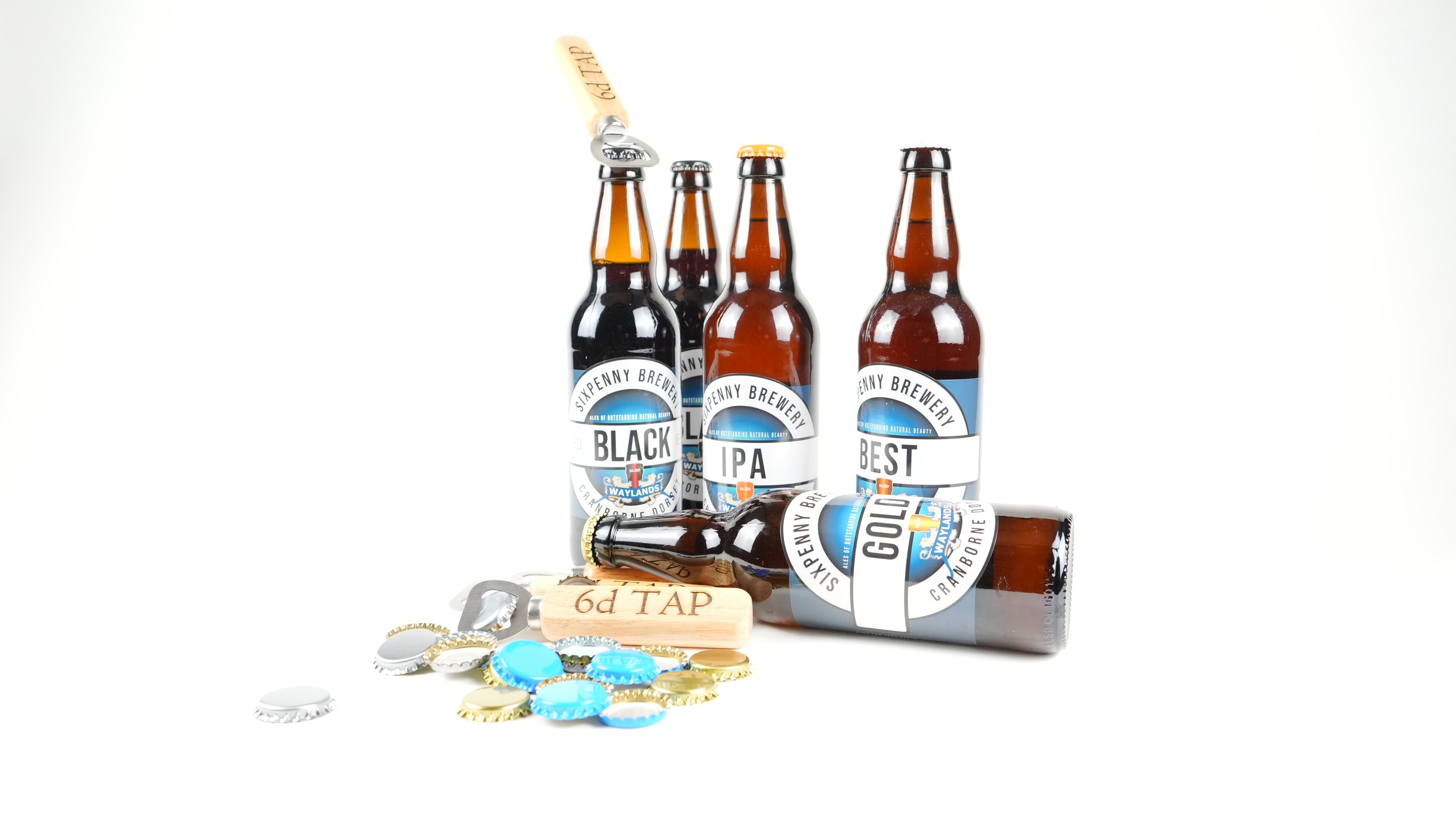 Four bottles of beer from SkyPenny Brewery, labeled Black, IPA, Best, and Gold, with one lying on its side and two beer bottle caps, a bottle opener, and a wood tap handle labeled '6d TAP' in front of them, all on a white background.