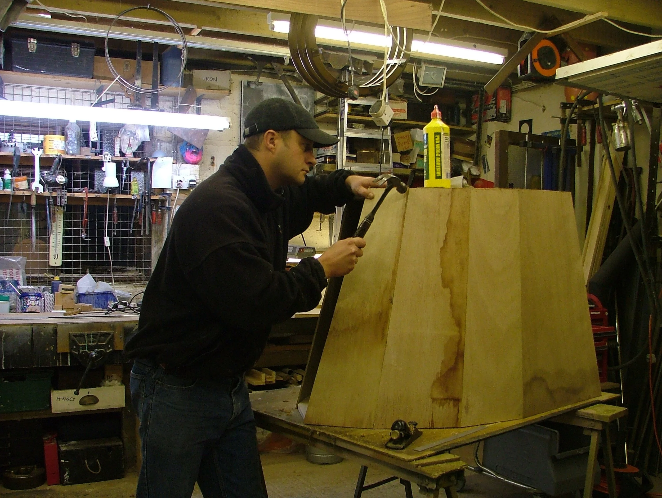 A man in a black jacket and cap is working in a woodworking shop, hammering a nail into a large wooden structure.