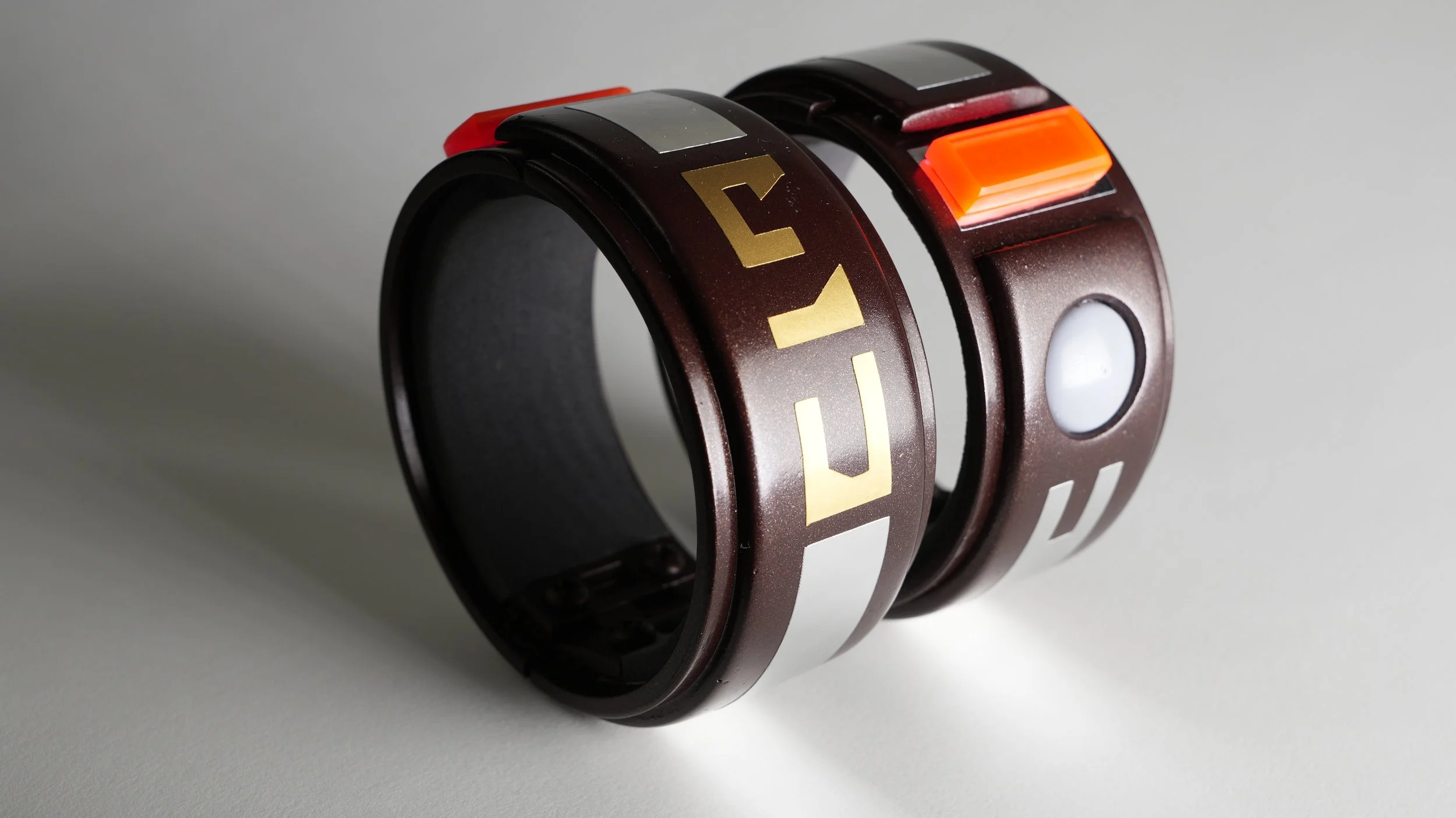 Two futuristic black wristbands with gold and white accents, one with the word 'HUB' and the other with various sensors and buttons, on a light gray surface.