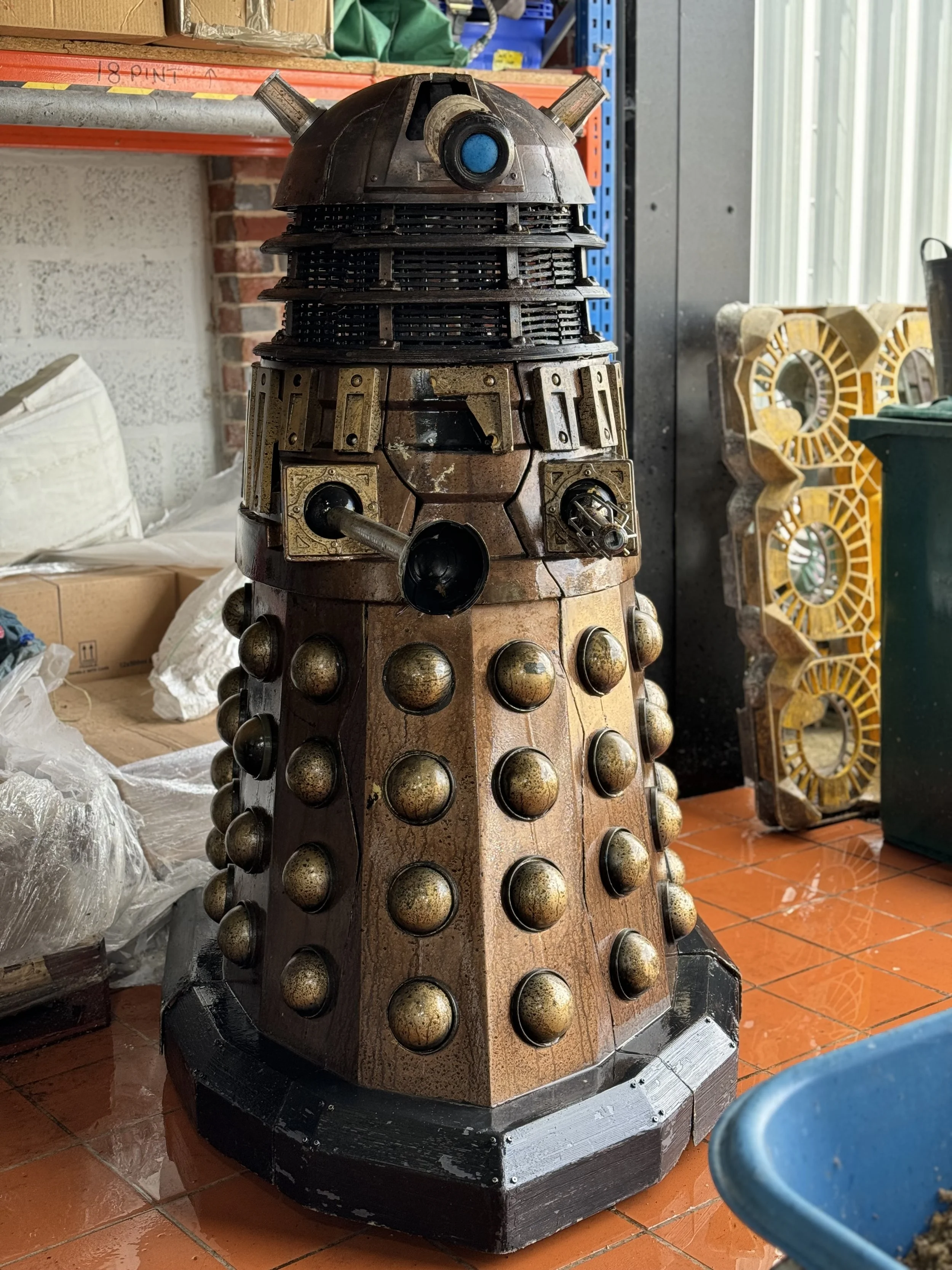 A detailed model or sculpture of a Dalek from Doctor Who, made from various metallic and plastic components, standing on a tiled floor and surrounded by boxes and industrial shelves.