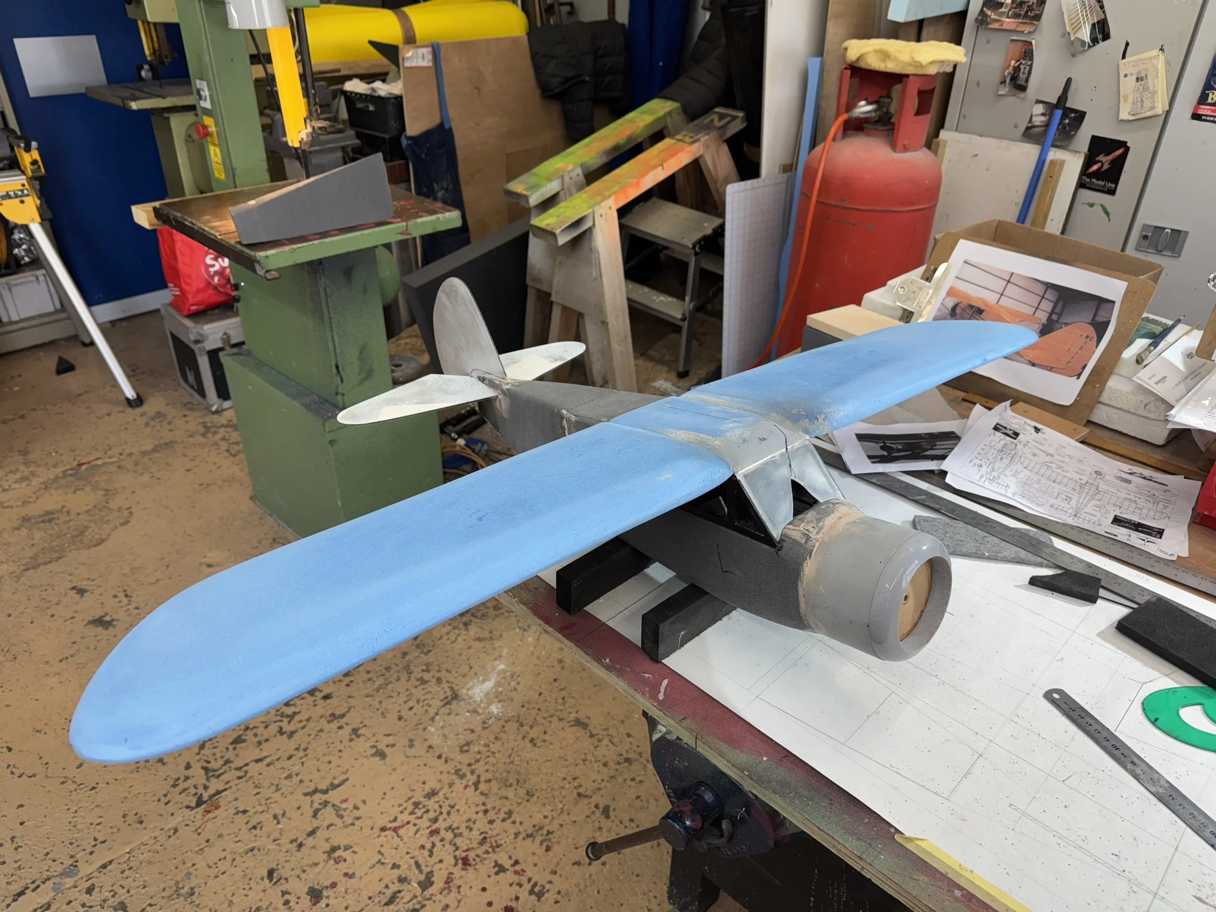 Model airplane with blue wings and silver tail on a workbench in a workshop surrounded by tools, plans, and equipment.