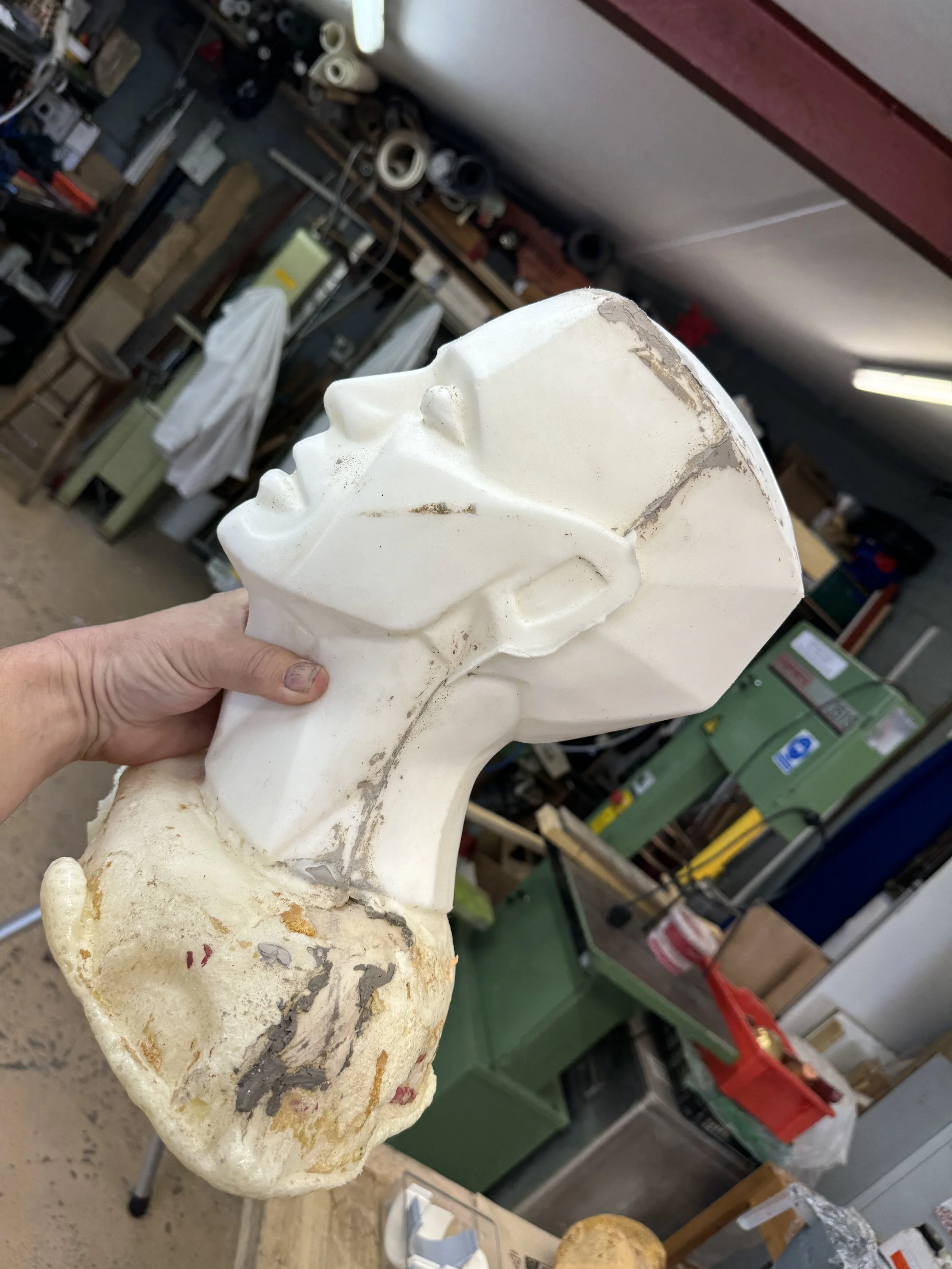A person holding a white plaster bust of a person's head and face in a workshop with various tools and equipment in the background.