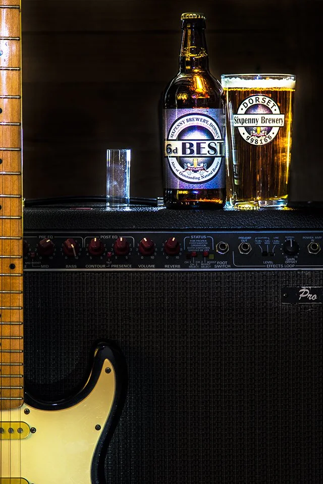 A glass of beer and a bottle of beer placed on top of an audio amplifier, with part of a guitar visible on the left side of the image.