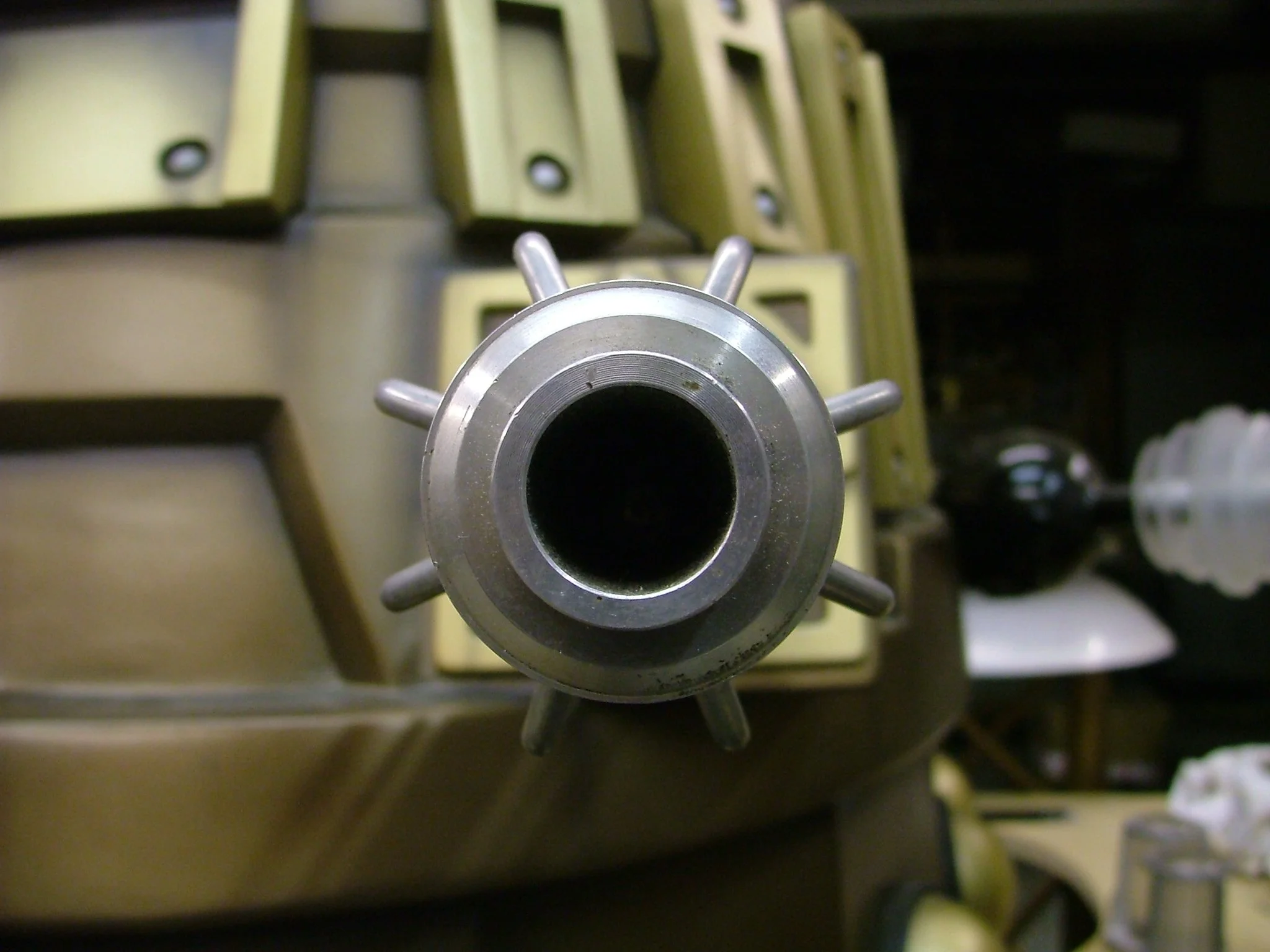 Close-up of a metal mechanical part with a central black opening and multiple protruding nozzles, in a workshop setting with blurred tools and equipment in the background.