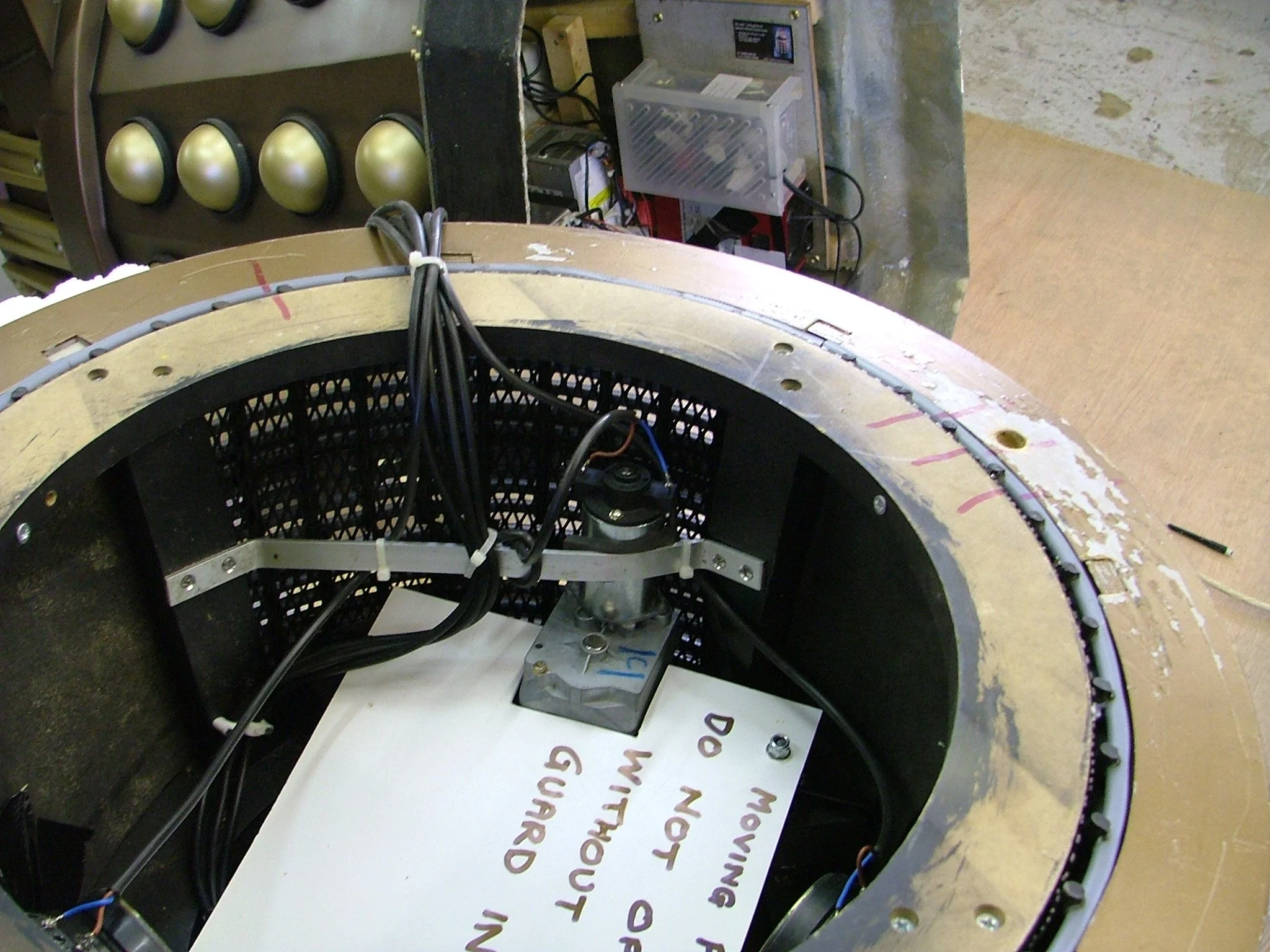 Close-up view of an electronic device with wires inside a circular frame, with a handwritten note placed beneath it. In the background, there are several spherical objects on a display and electronic equipment attached to a wall.