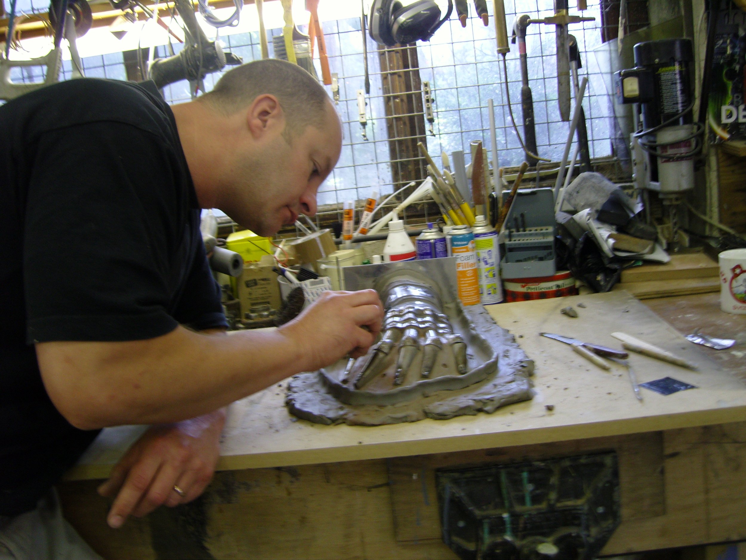 A man working on a sculpture or model in a workshop filled with tools, supplies, and a window in the background.