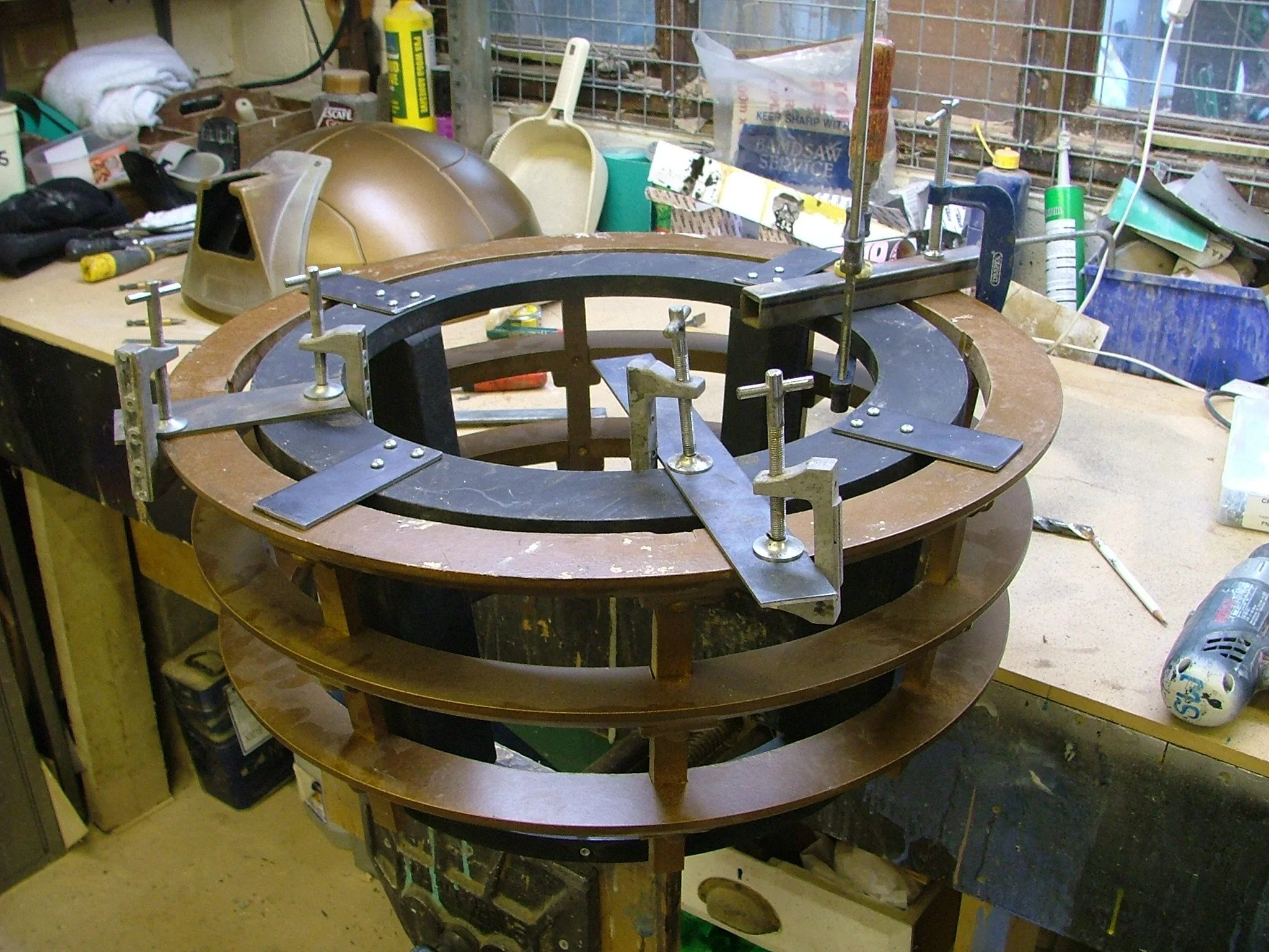 A woodworking project in progress on a workbench, with circular wooden and metal components clamped and assembled for construction.