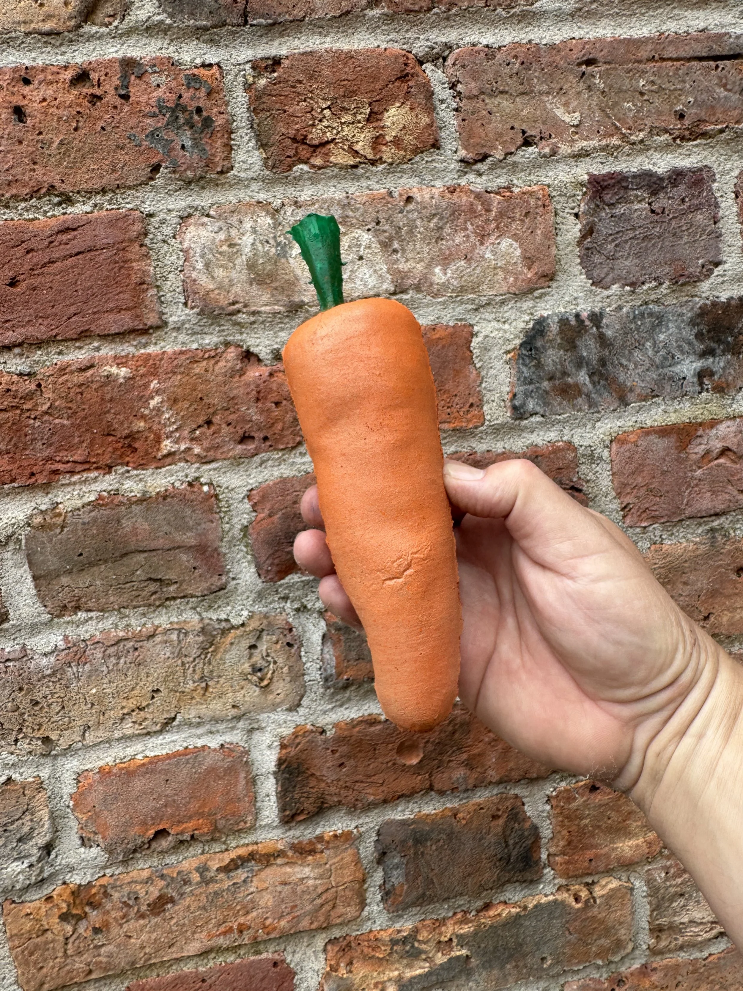 A hand holding a plush toy shaped like a carrot, with an orange body and green top, against a brick wall background.