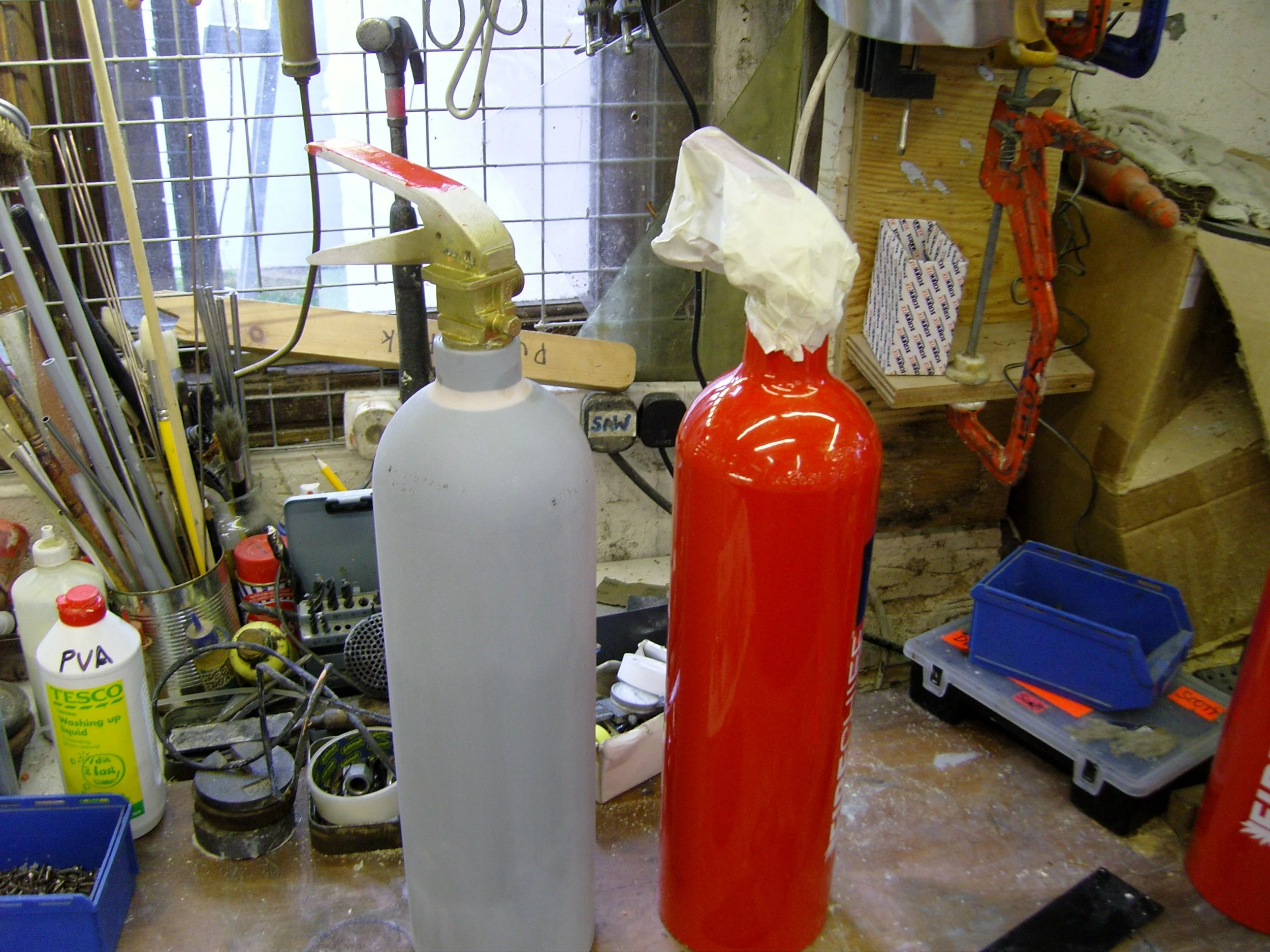 In a workshop, there are two fire extinguishers, one white and one red, placed on a workbench surrounded by various tools and materials.