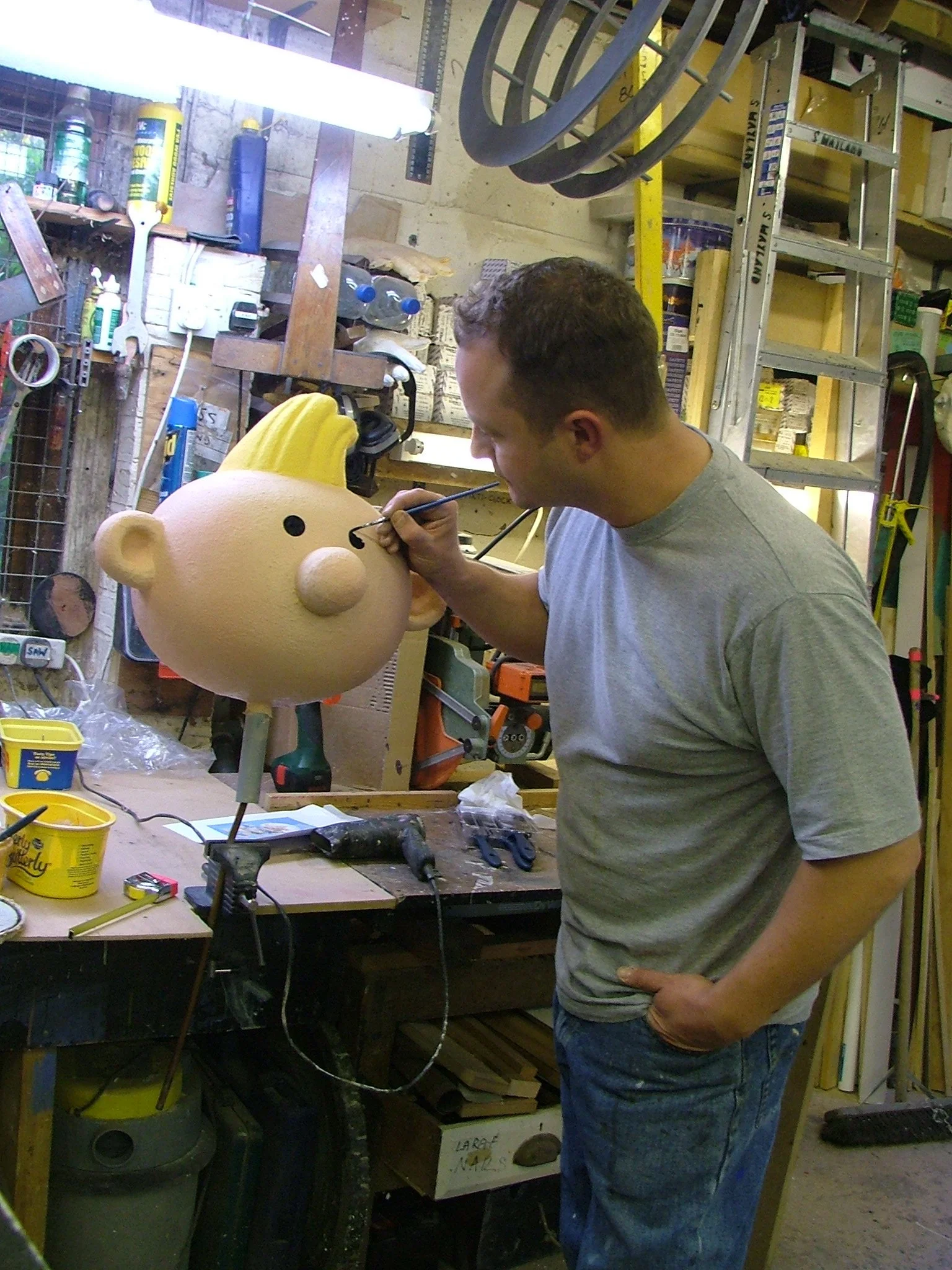 A man painting a cartoonish large head sculpture of a person with a yellow mohawk, in a workshop filled with tools and supplies.