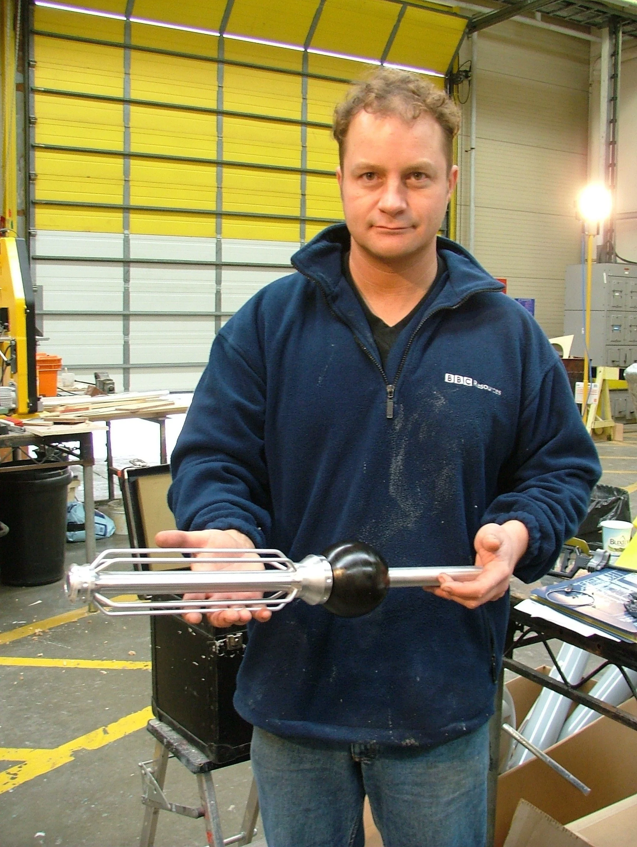 A man holding a metal antenna with a large black ball at one end inside a workshop or industrial space.