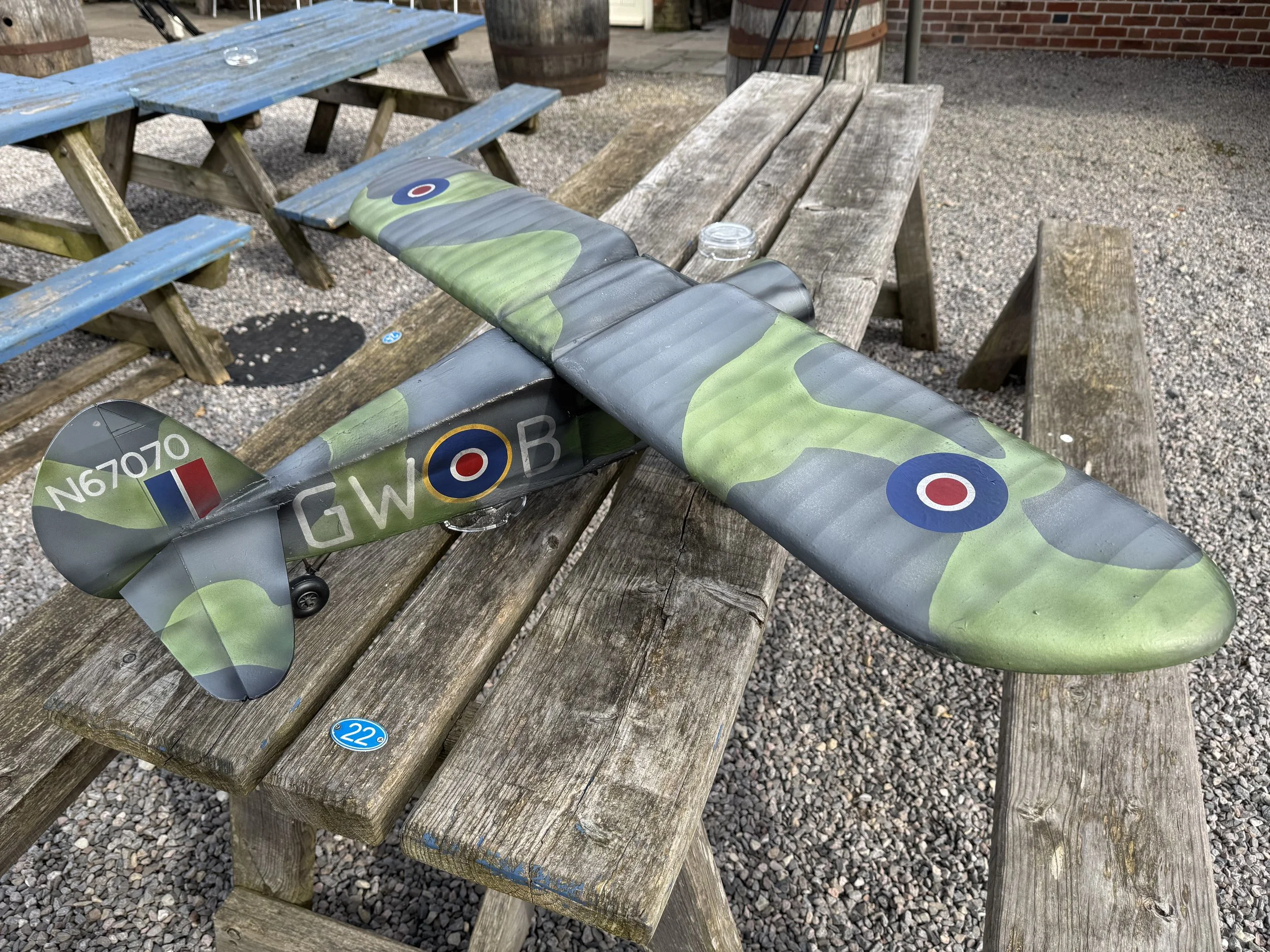 A model airplane with a green camouflage pattern and British Royal Air Force roundel markings on the wings and fuselage, resting on a weathered wooden picnic table outdoors.