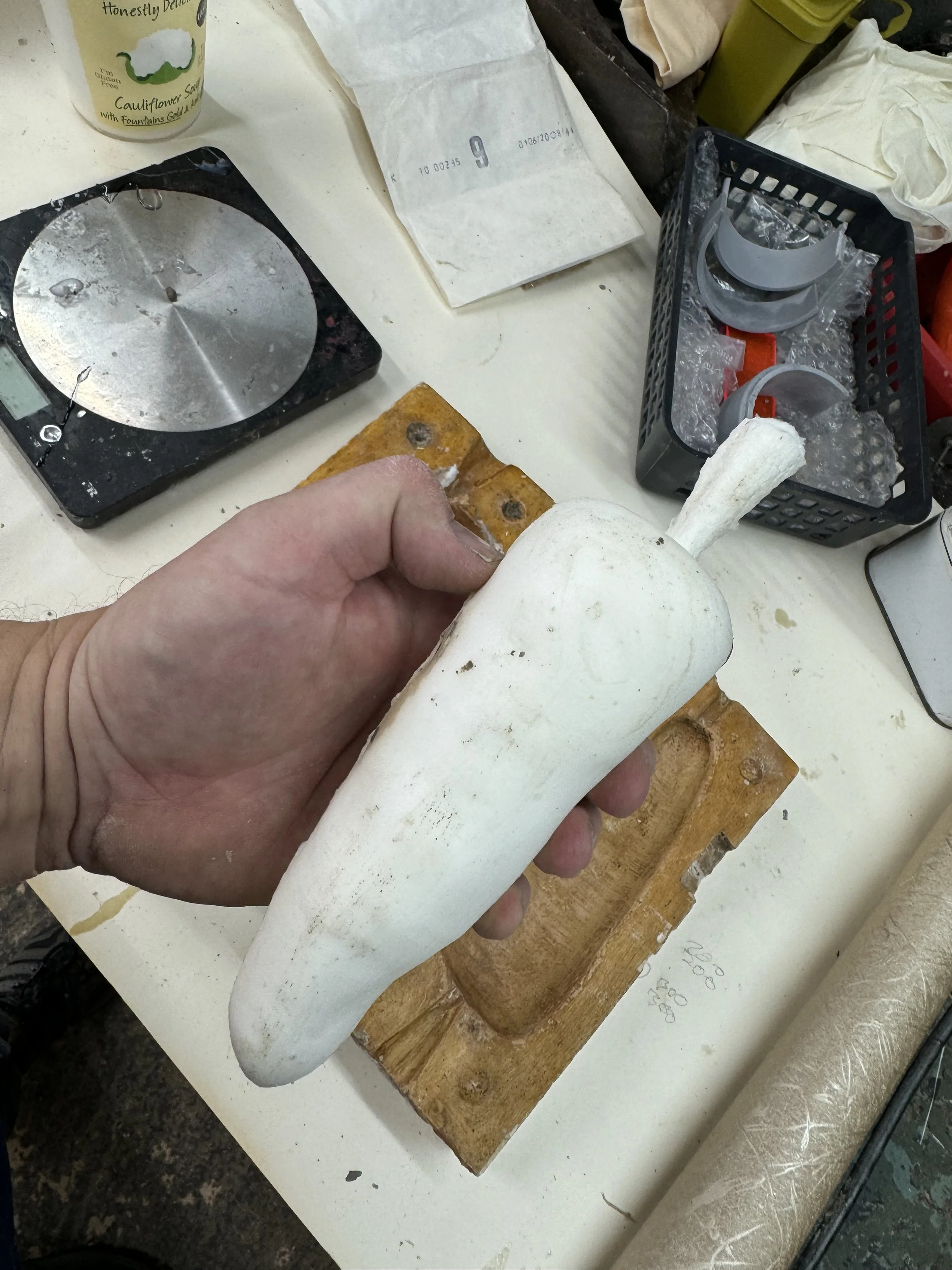A person holding a large white radish on a wooden cutting board. Various kitchen items and a paper bag are visible on the table, including a scale, a basket with plastic lids, a cauliflower seed packet, and a paper roll.