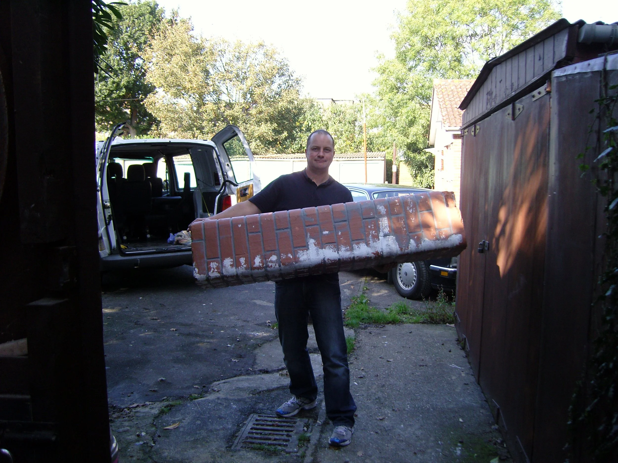 A man holding a brick appears to be in a driveway or backyard next to a van with open sliding door, a wooden shed, and trees in the background.