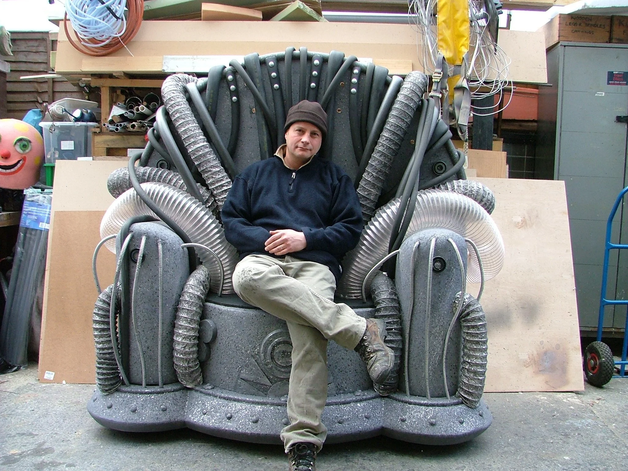 A man sitting on a sci-fi styled chair made of various pipes and industrial materials in a workshop or storage area.