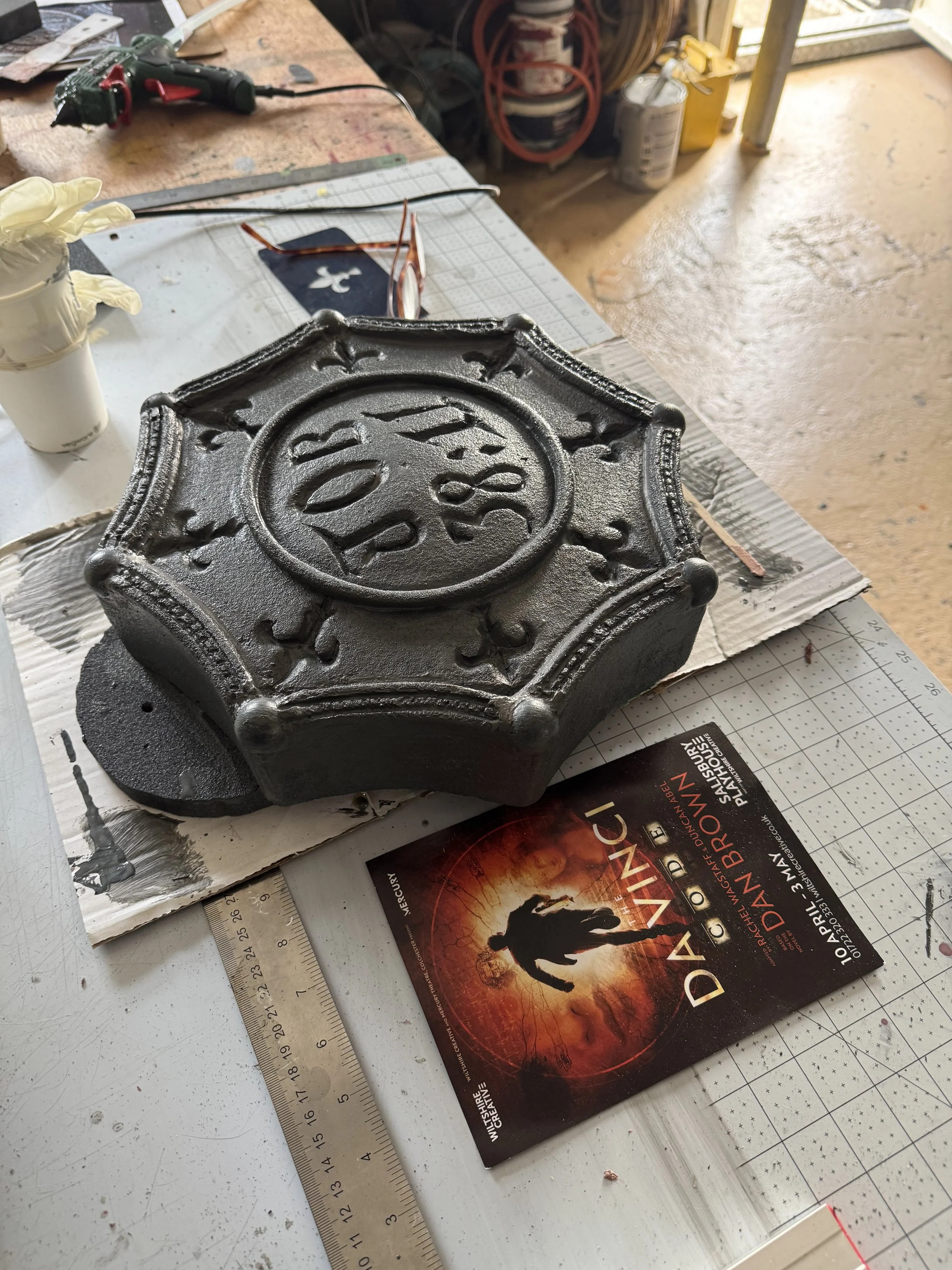 Metallic sheriff badge sculpture resting on a work table, with tools, a ruler, and a 'Day of the Dead' promotional card nearby in a mechanic or artist's workshop.