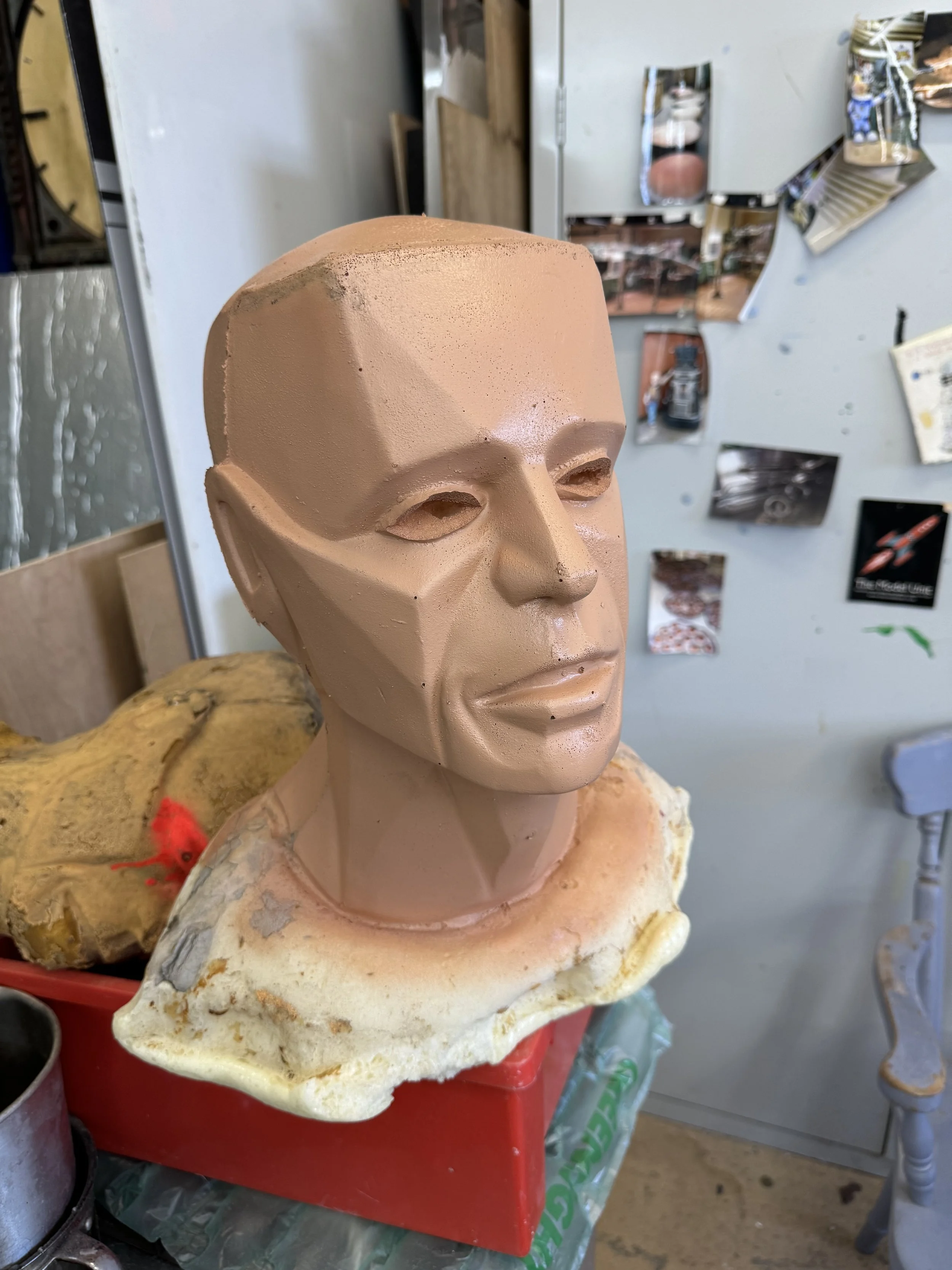 A clay sculpture of a human head with detailed facial features, resting on a piece of foam or foam-like material in an artist's studio with photos and references on the wall.