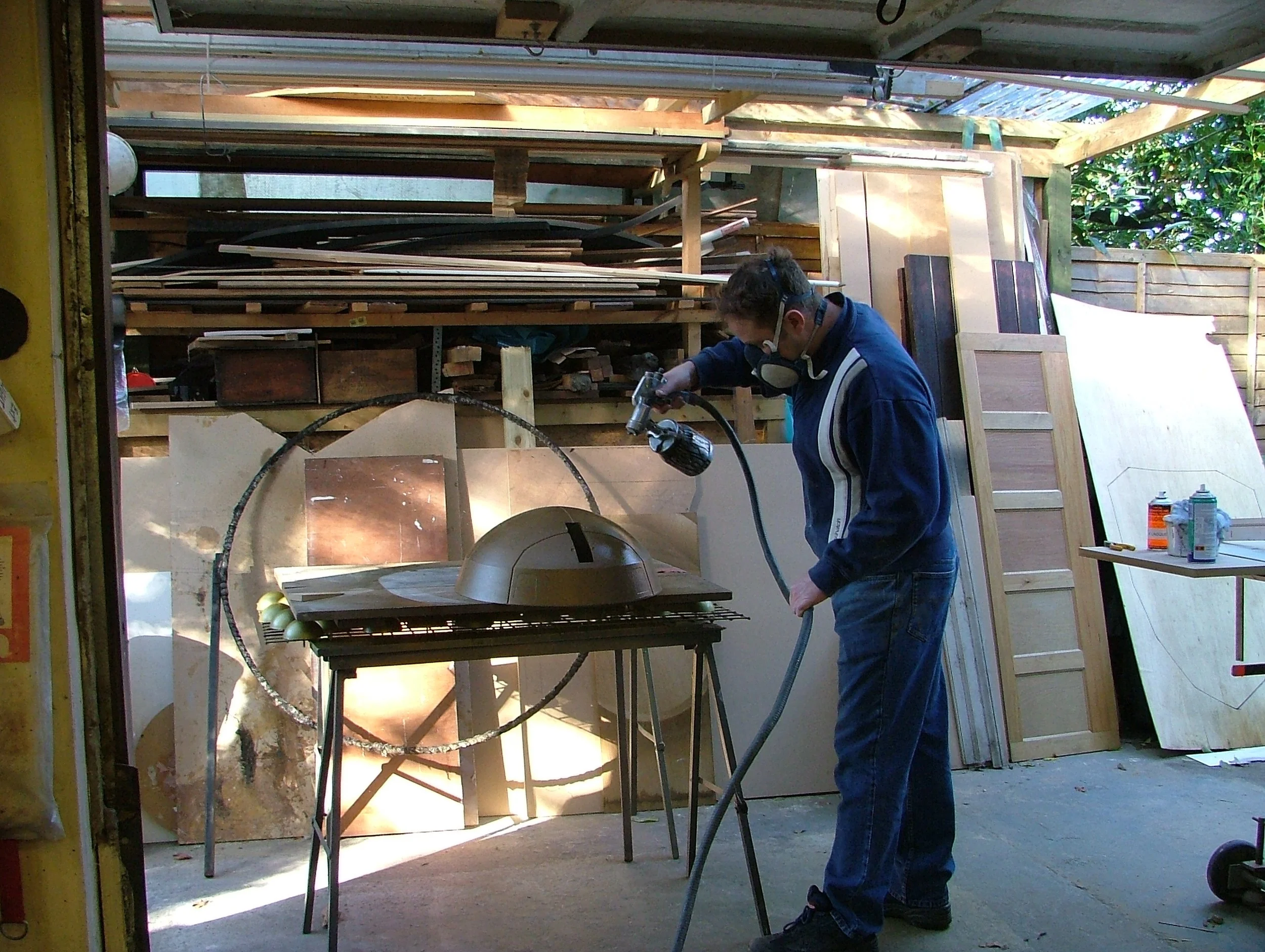 A person wearing safety goggles and a face mask working with a spray tool in a woodworking shop, with various wooden boards and tools around.