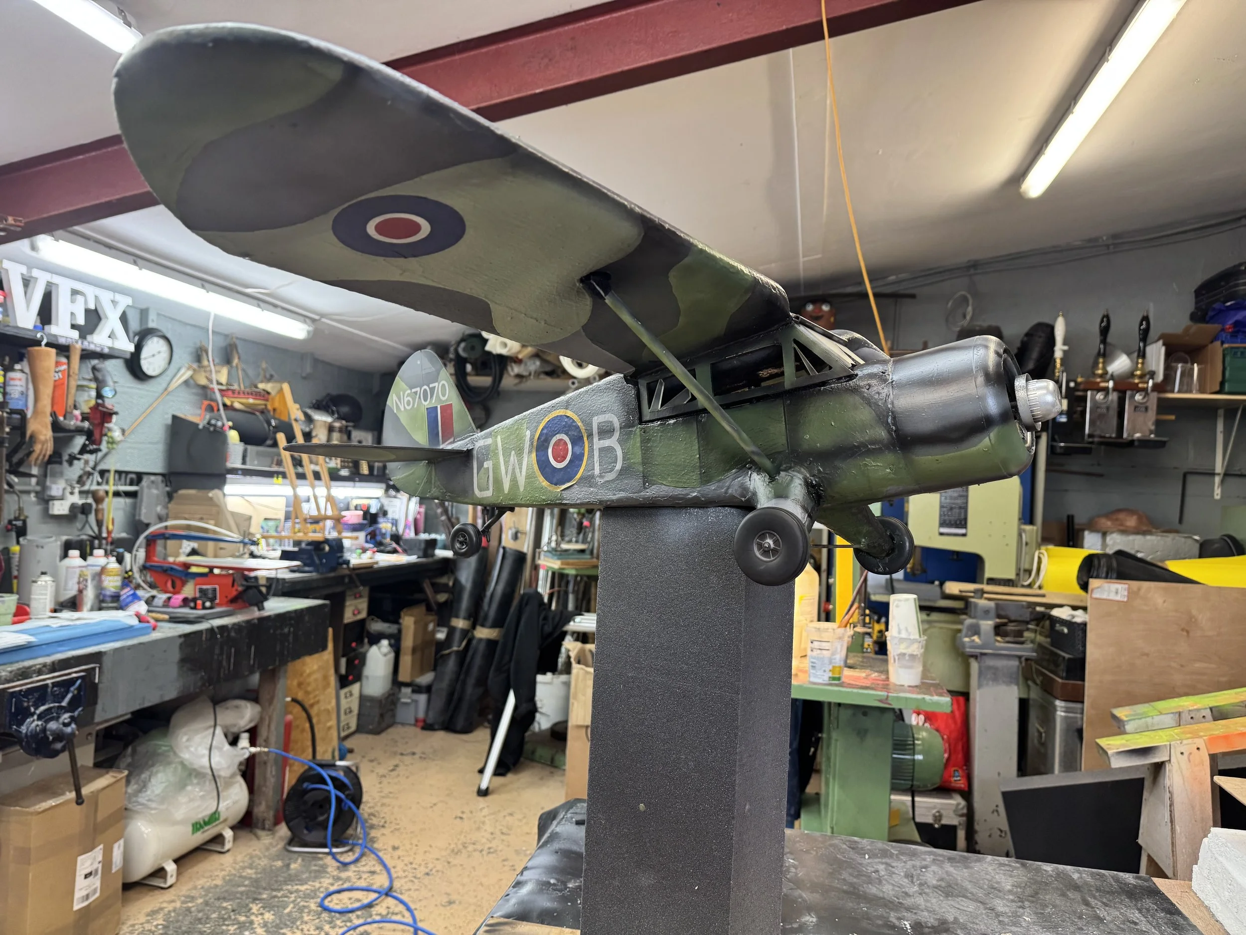 Model of a vintage military aircraft with camouflage paint, displayed on a stand inside a cluttered workshop.