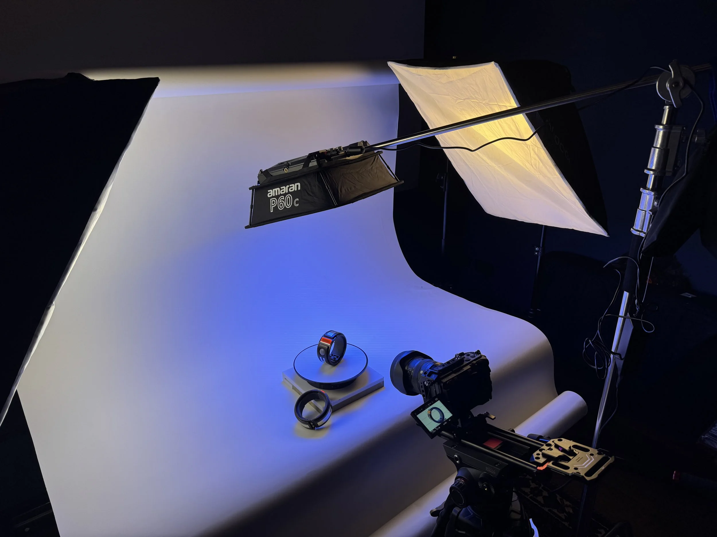 Photography studio setup with lighting equipment, a camera on a tripod, a white seamless backdrop, and a circular light with a band on it, arranged on a white table.