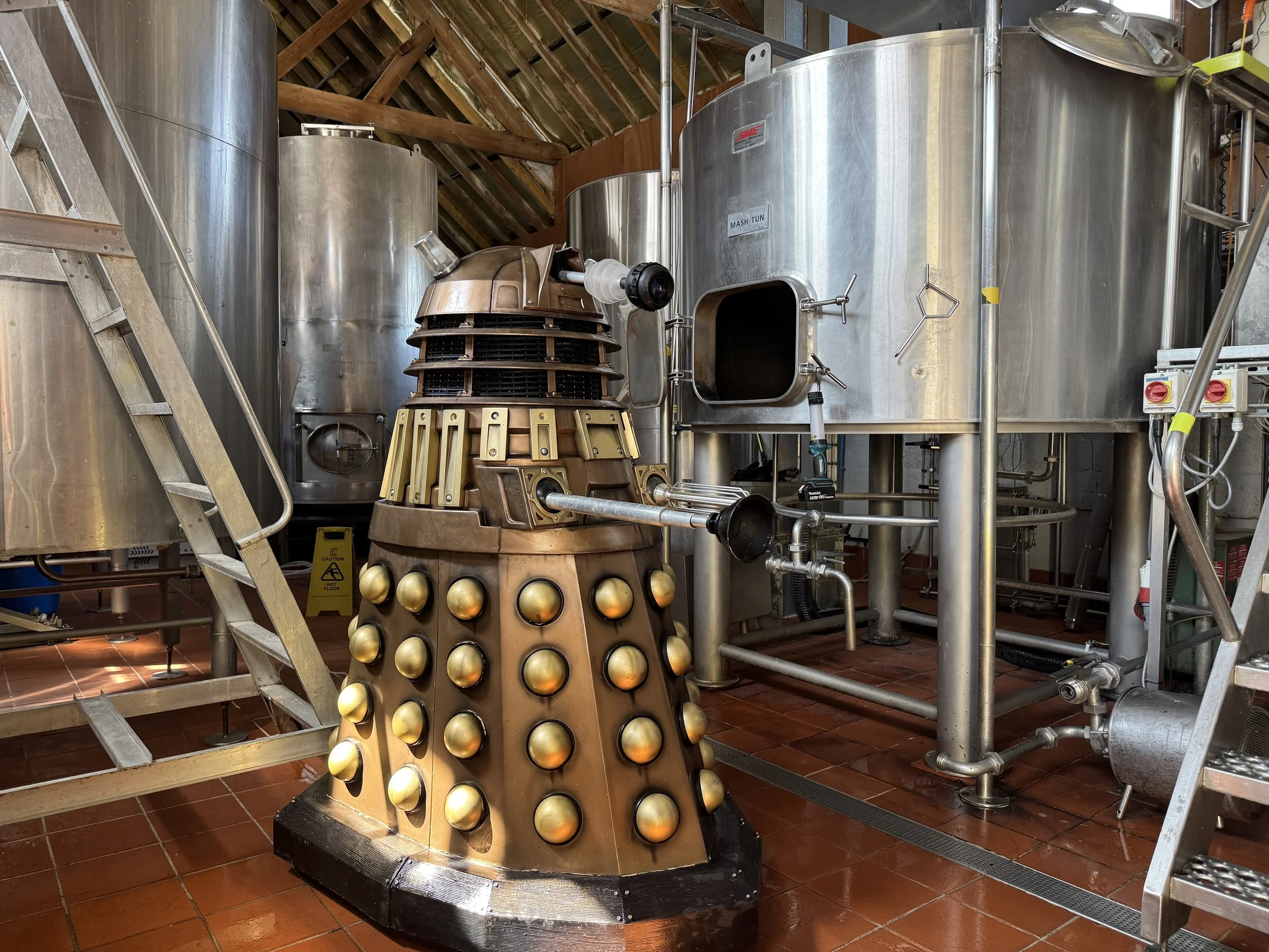 A life-size replica of a Dalek from Doctor Who in an industrial setting with metal tanks, pipes, and a wooden ladder in the background.
