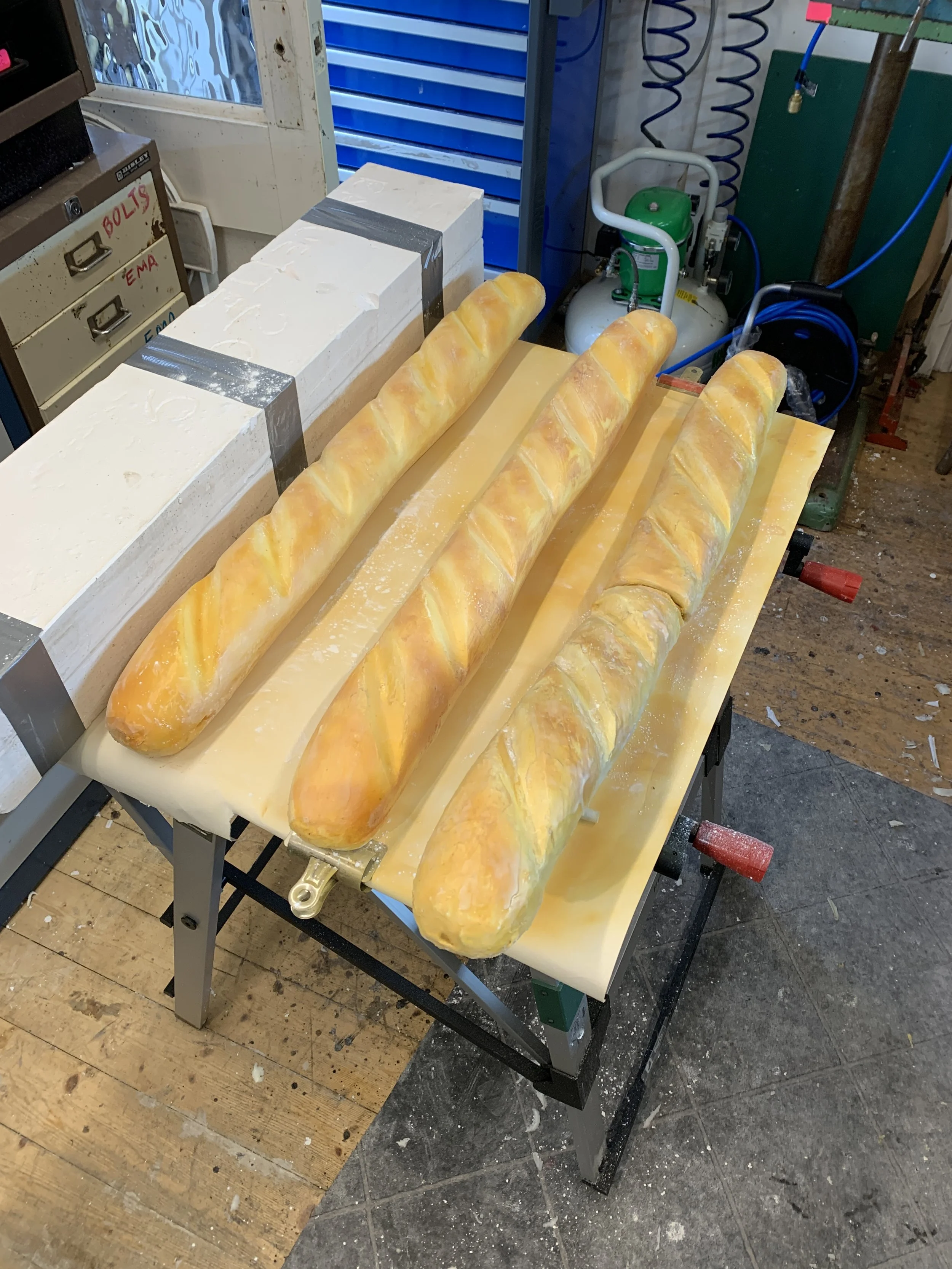 Three golden baguettes on a yellow cutting board in a bakery or kitchen setting with baking supplies and equipment visible around.