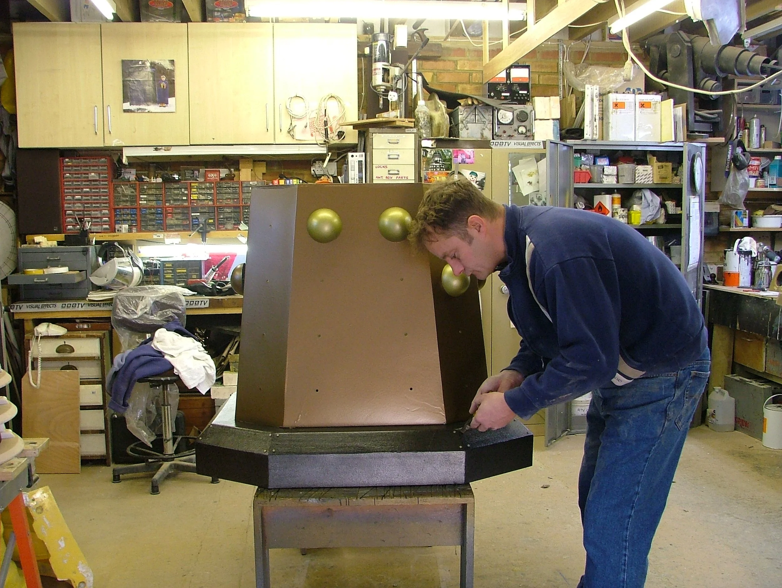 Man working on a wooden structure with gold spheres in a cluttered workshop with tools, shelves, and cabinets.