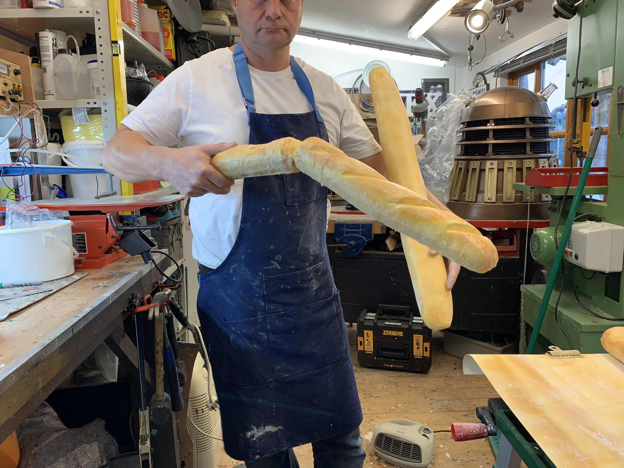 A man holding three large baguettes in a workshop or bakery setting with various tools and equipment around him.