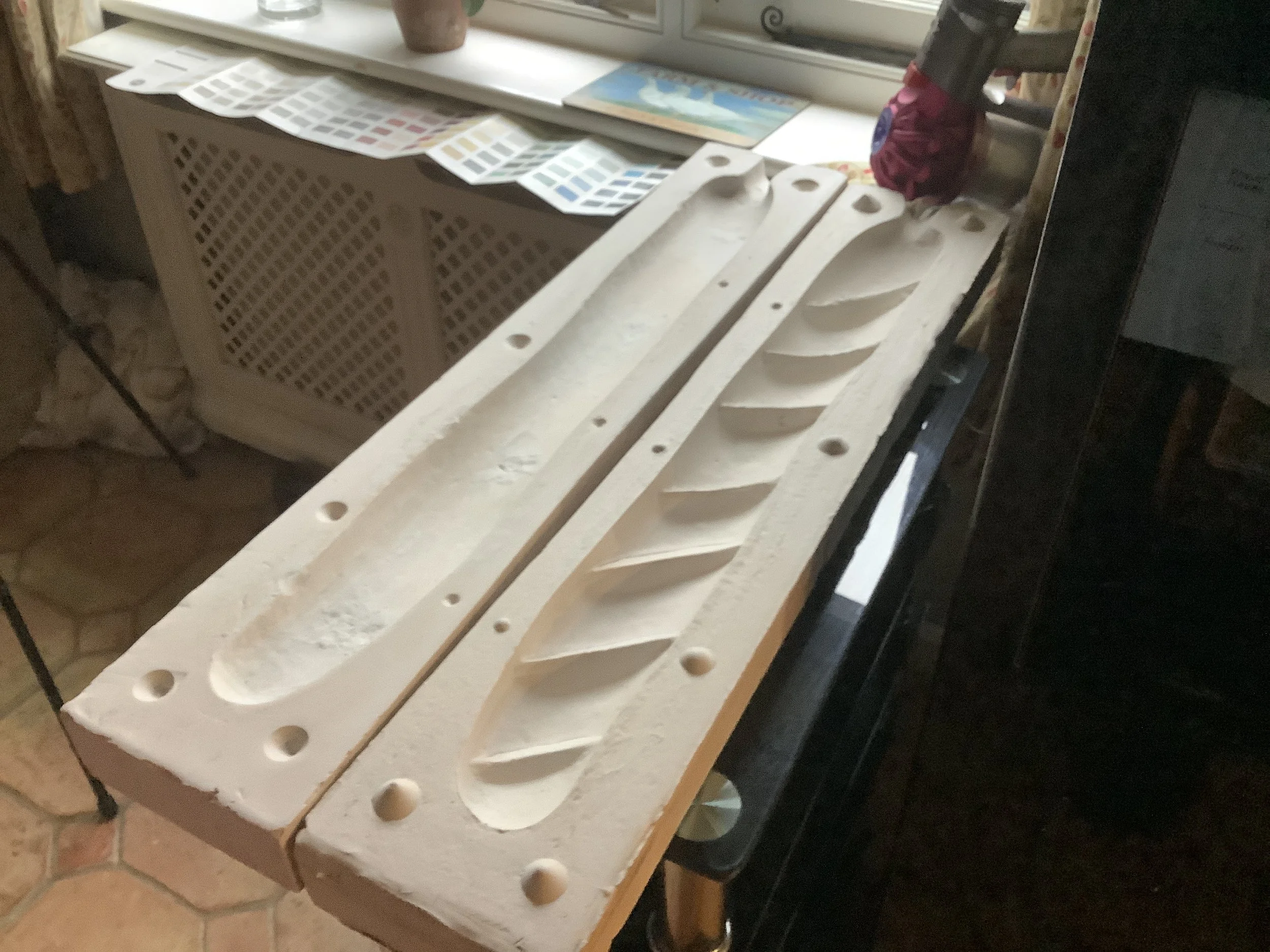 A worktable with carved plaster molds or sculptures, featuring elongated, curved indentations and circular holes, set in a room with a window and a countertop with paint samples or color swatches.